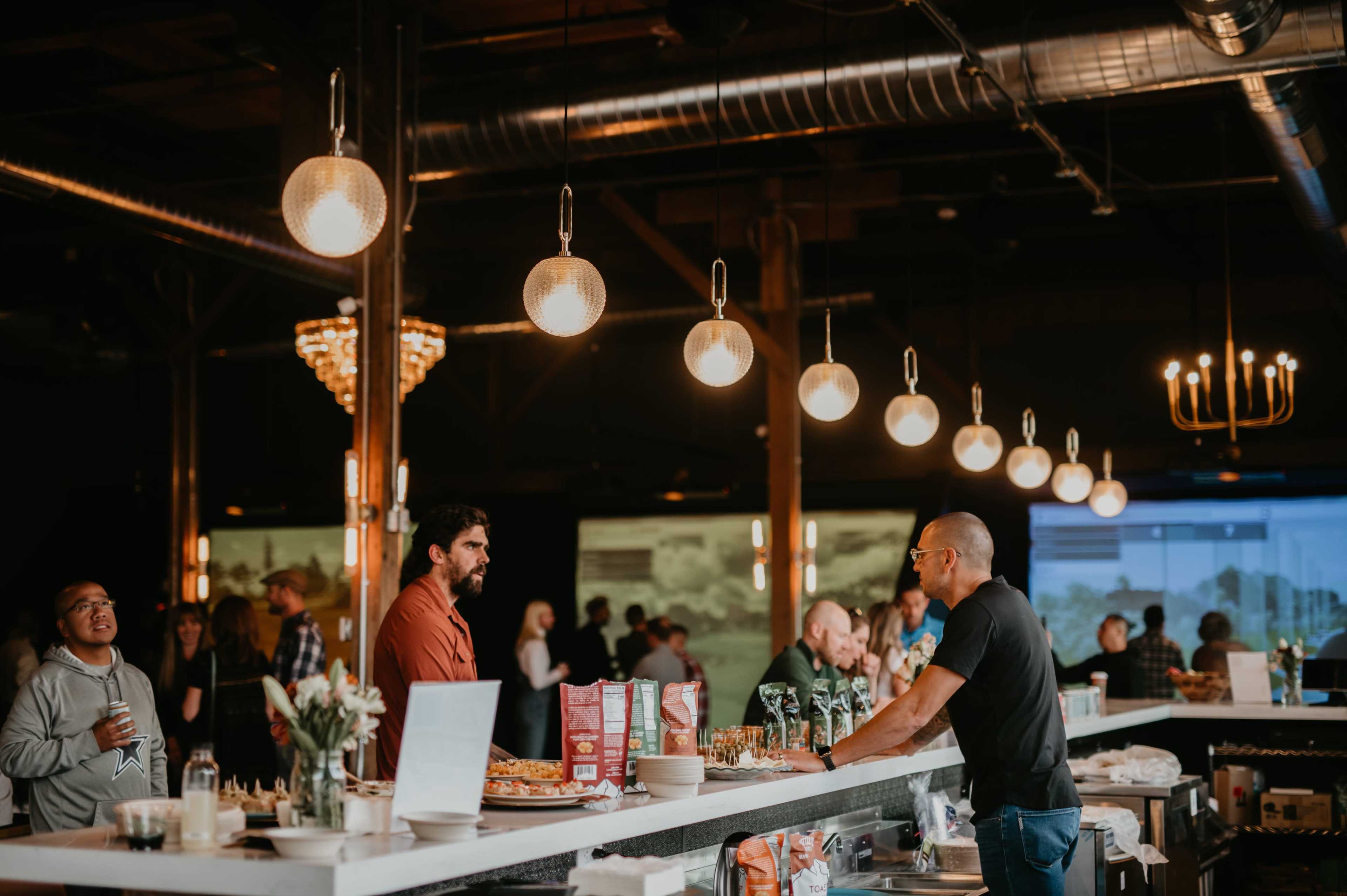 A lively bar scene features people interacting while a bartender serves drinks under pendant lights.
