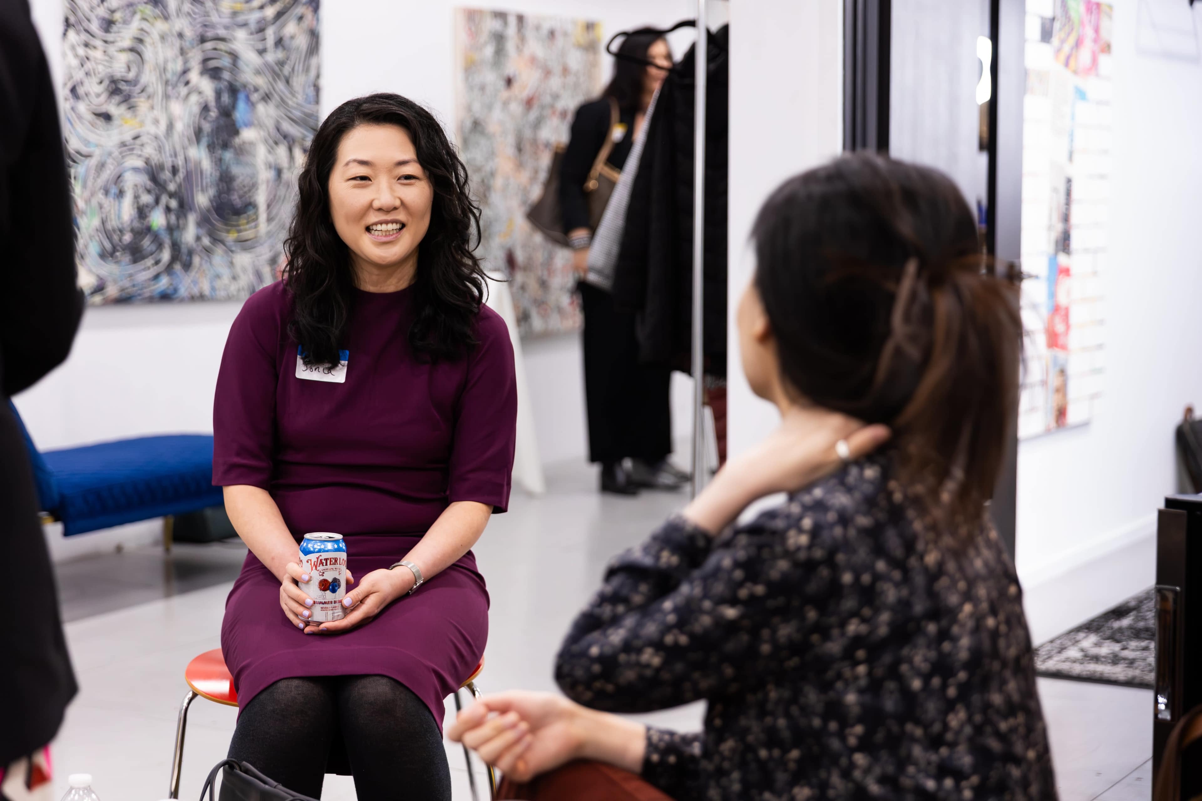 A woman in a purple dress holds a can while smiling and conversing with another person in a casual setting.