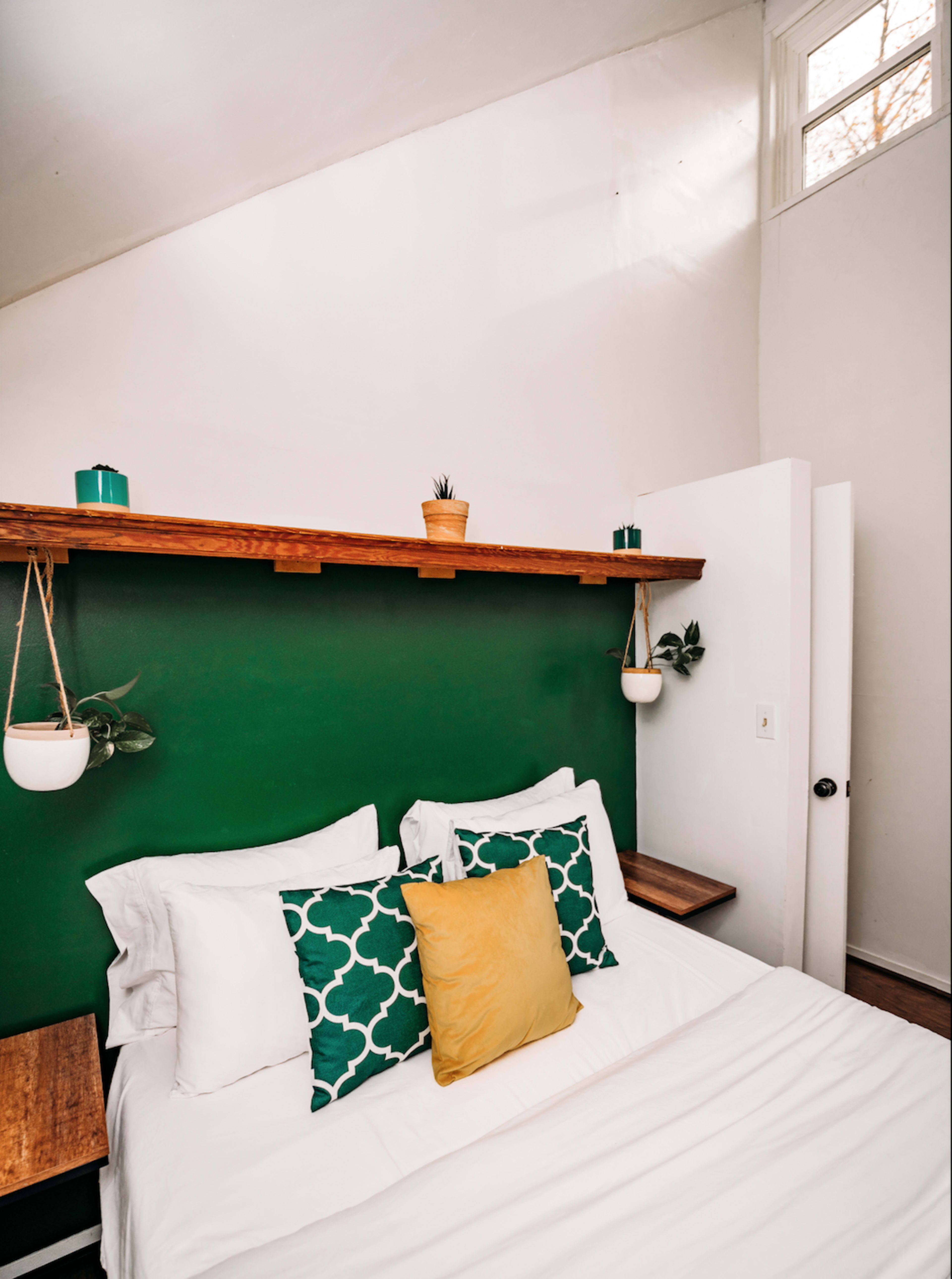 The image shows a neatly made bed with white linens and decorative pillows against a green accent wall, topped by a wooden shelf with plants and small teal containers.