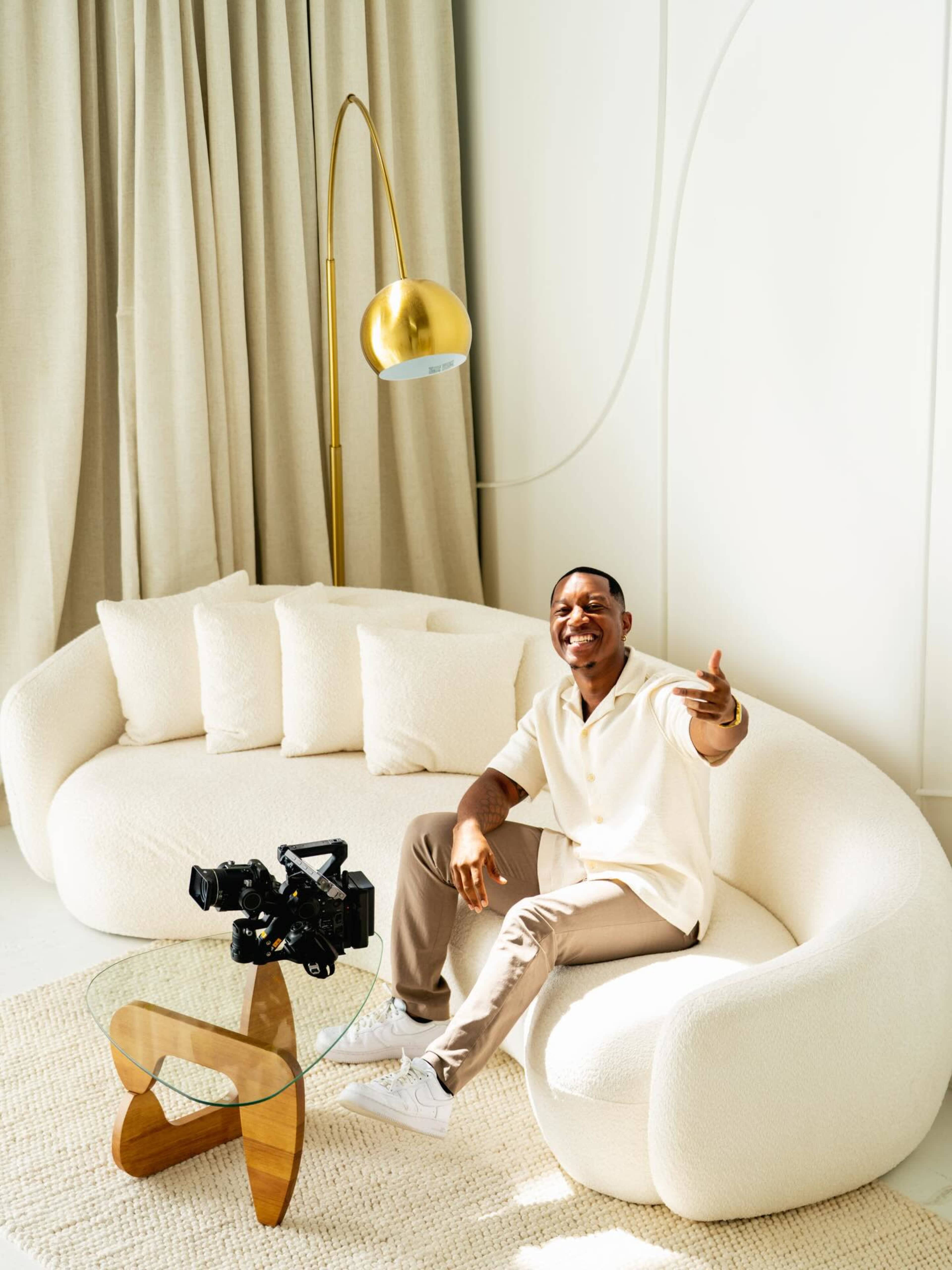 A person sits on a curved white sofa, smiling and gesturing, with a camera placed on a glass table nearby.