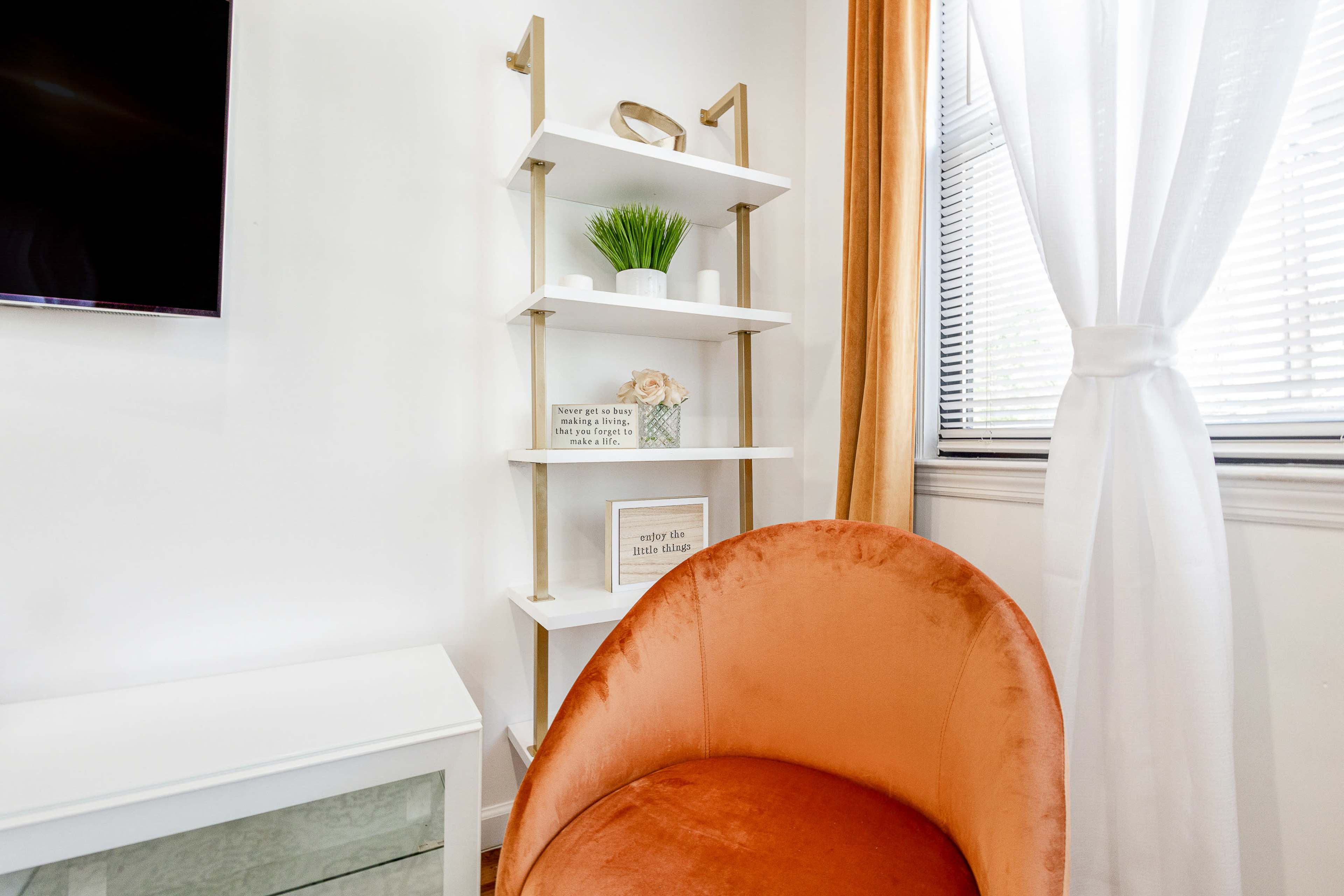A stylish living space with a velvet orange chair, a white shelving unit with decorative items, and a window adorned with sheer curtains.