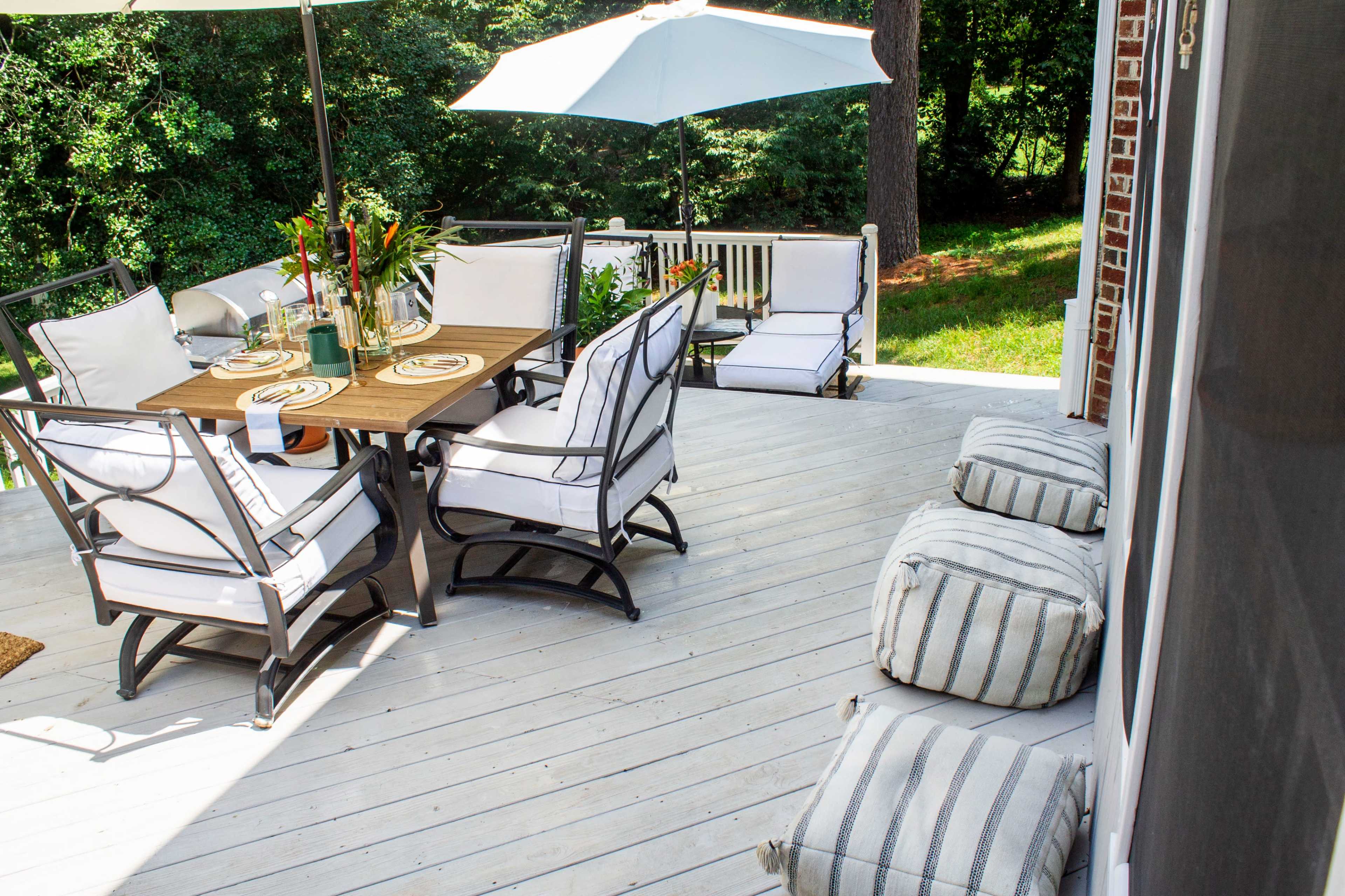 A wooden patio features a dining table with chairs, an umbrella, and additional seating on a deck surrounded by trees.