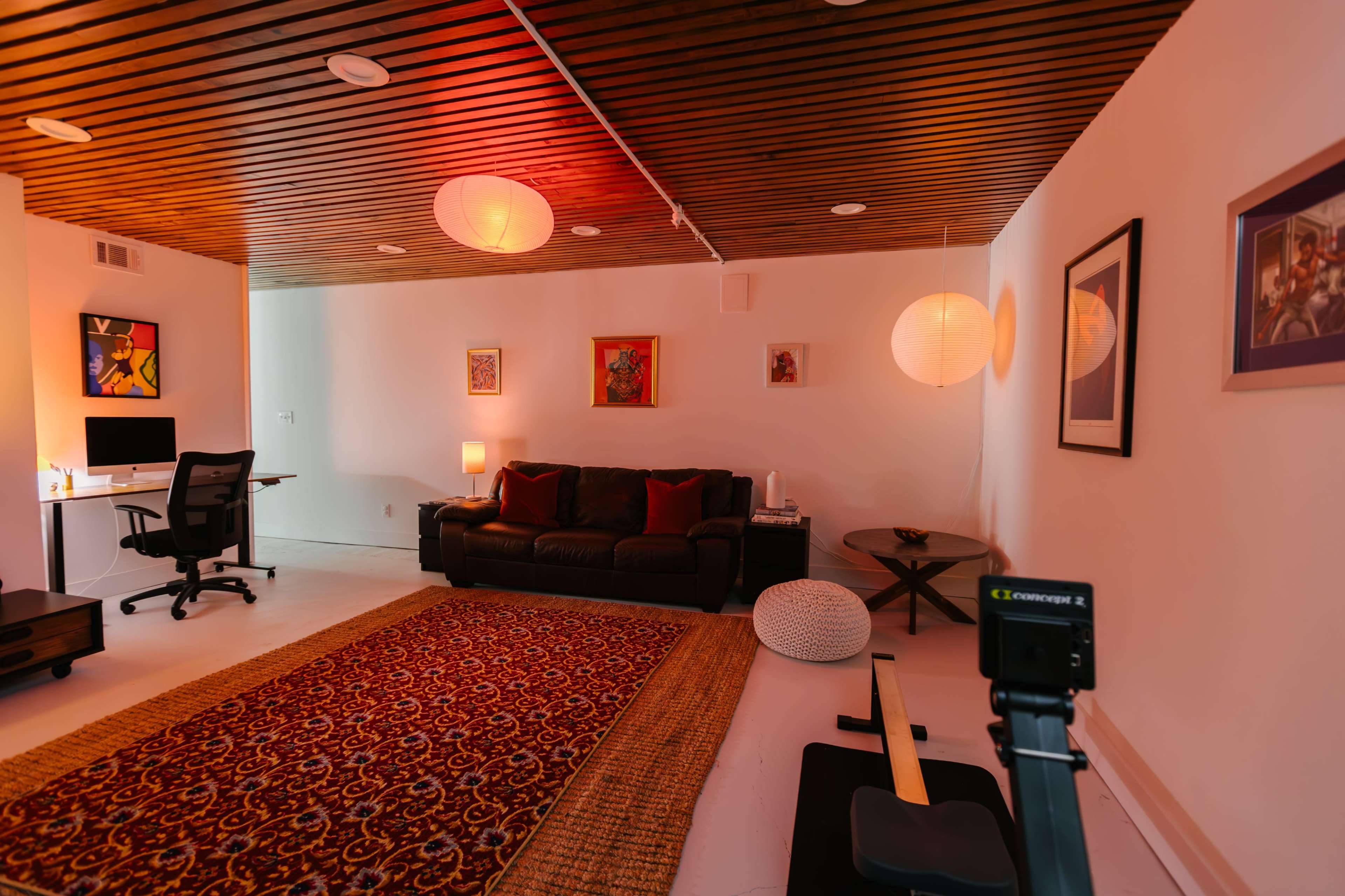 The image shows a spacious room featuring a brown couch, a wooden ceiling, a decorative rug, a desk with a computer, and soft lighting from paper lanterns.