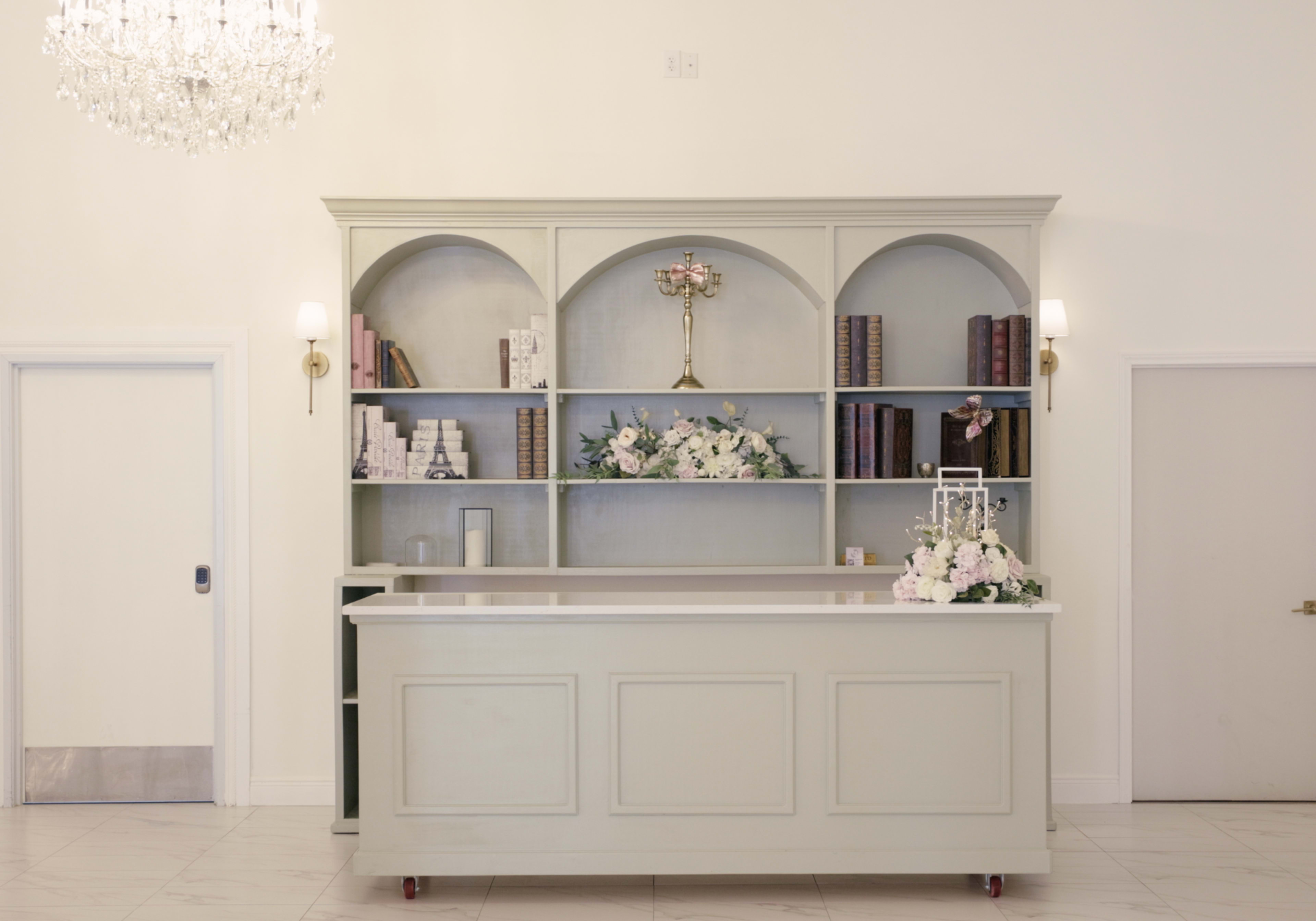The image shows a light-green reception desk with an arched shelving unit displaying books and floral arrangements, set against a plain white wall.