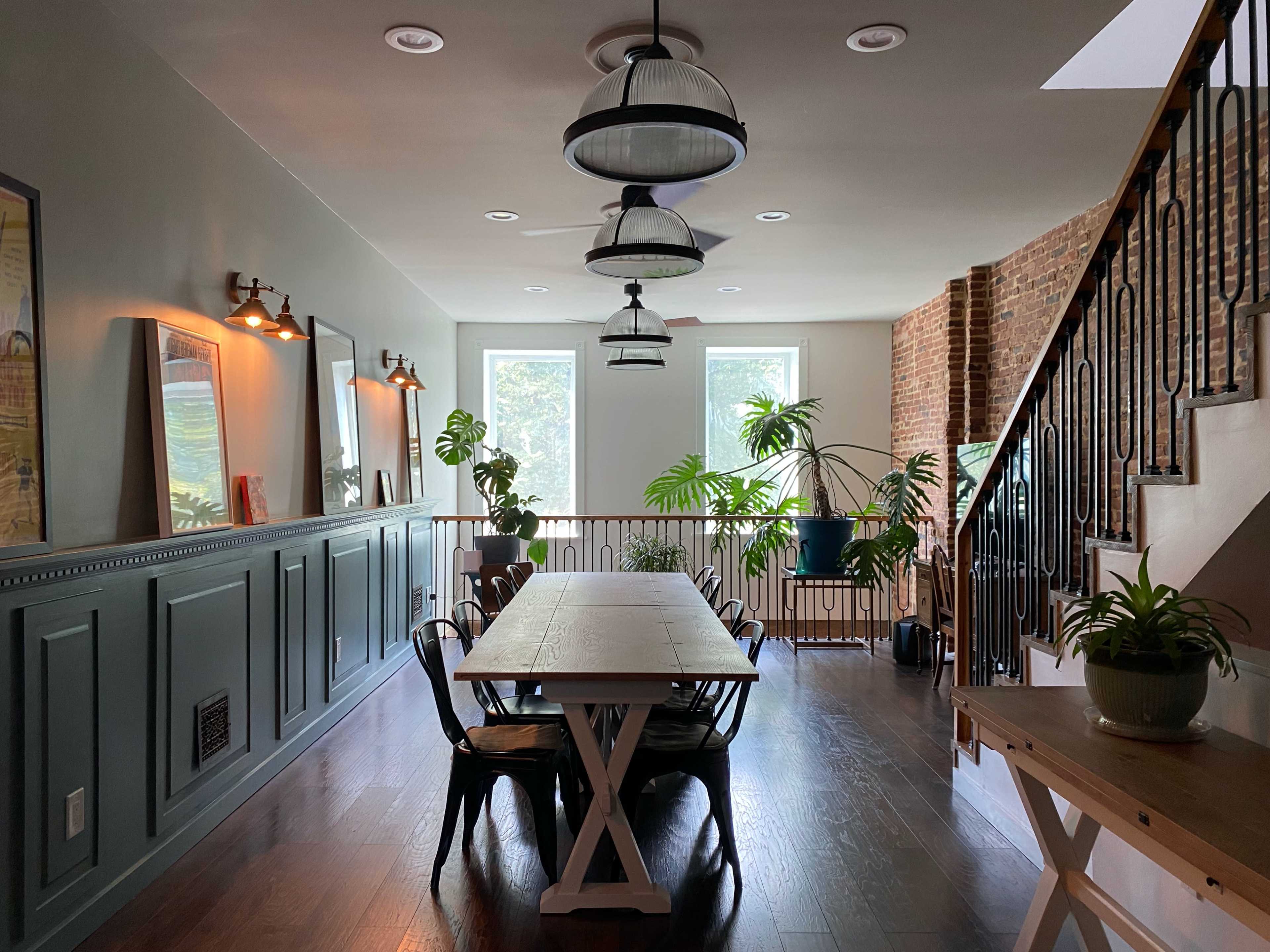 A dining area features a long wooden table surrounded by black chairs, with large windows allowing natural light and indoor plants decorating the space.