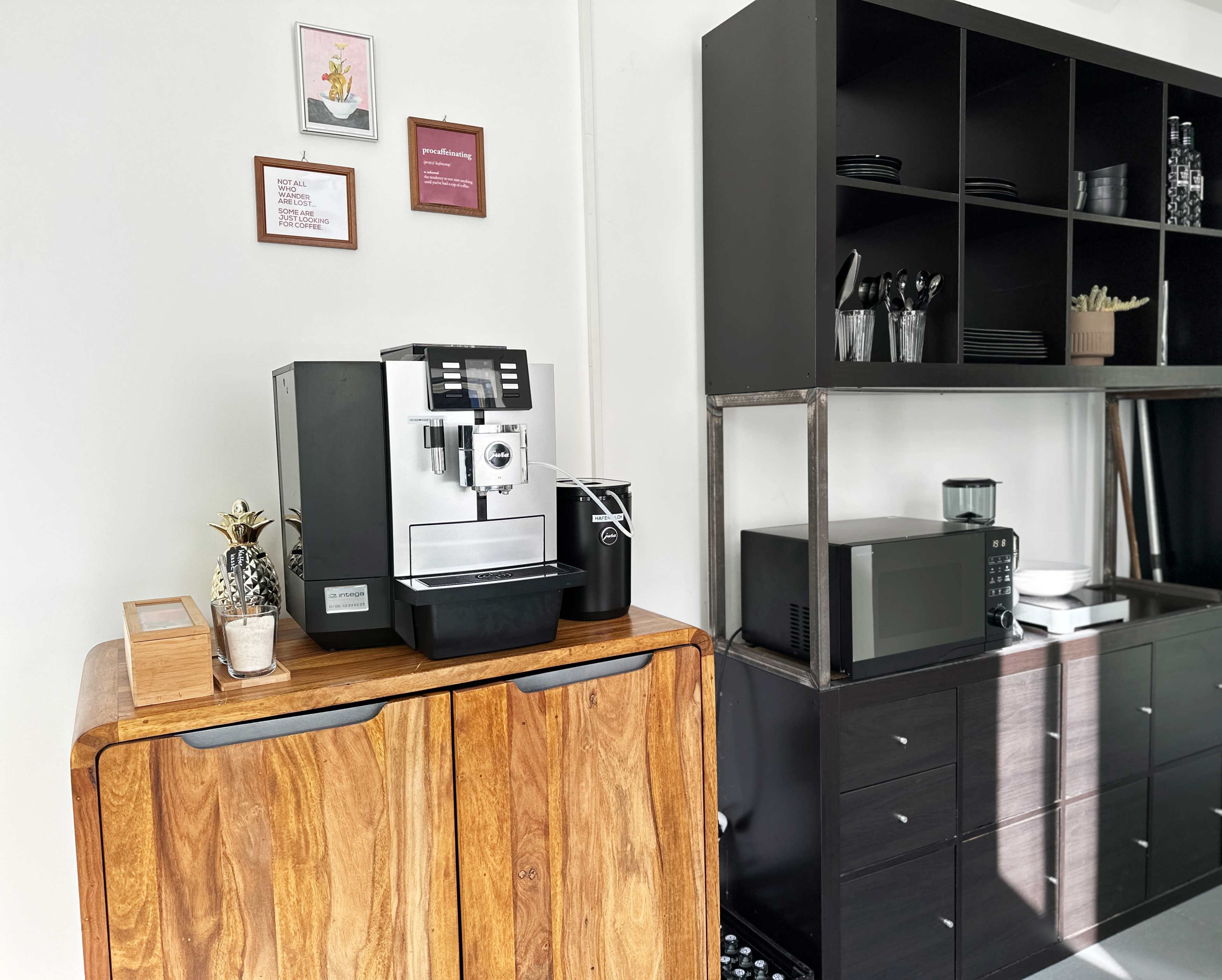 A modern kitchen corner featuring a coffee machine on a wooden cabinet, a microwave, and shelves holding glassware and utensils.