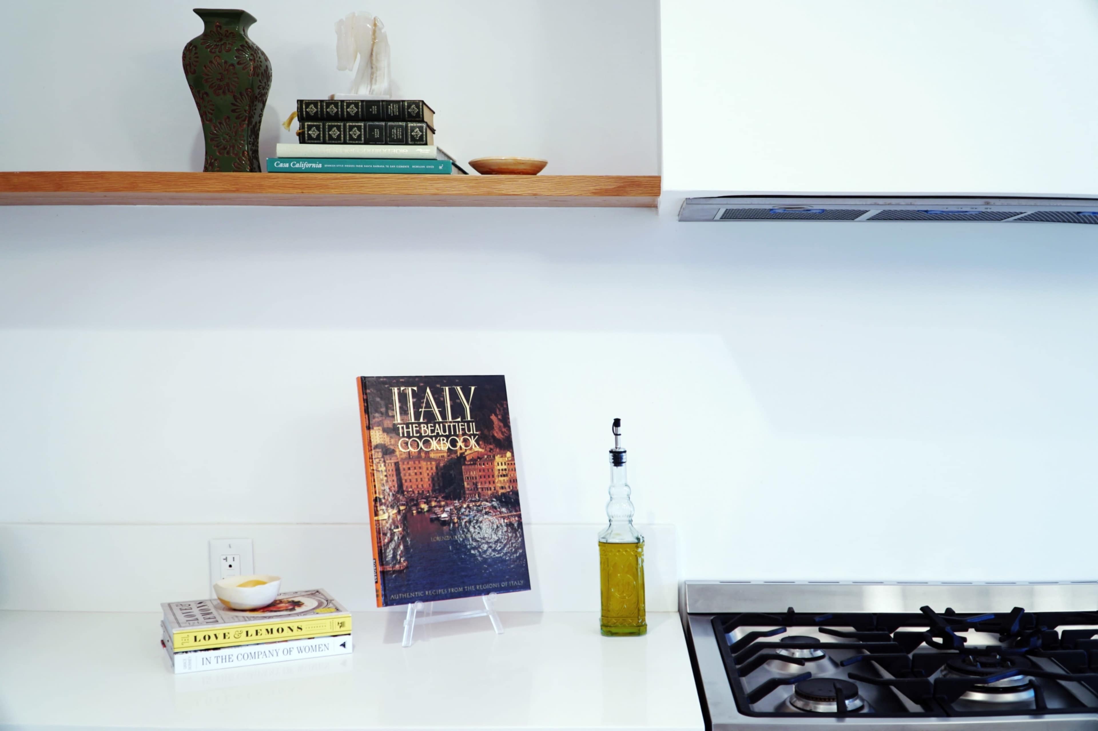 A modern kitchen shelf displays a cookbook titled "Italy: The Beautiful Cookbook," alongside decorative items and a bottle of olive oil.