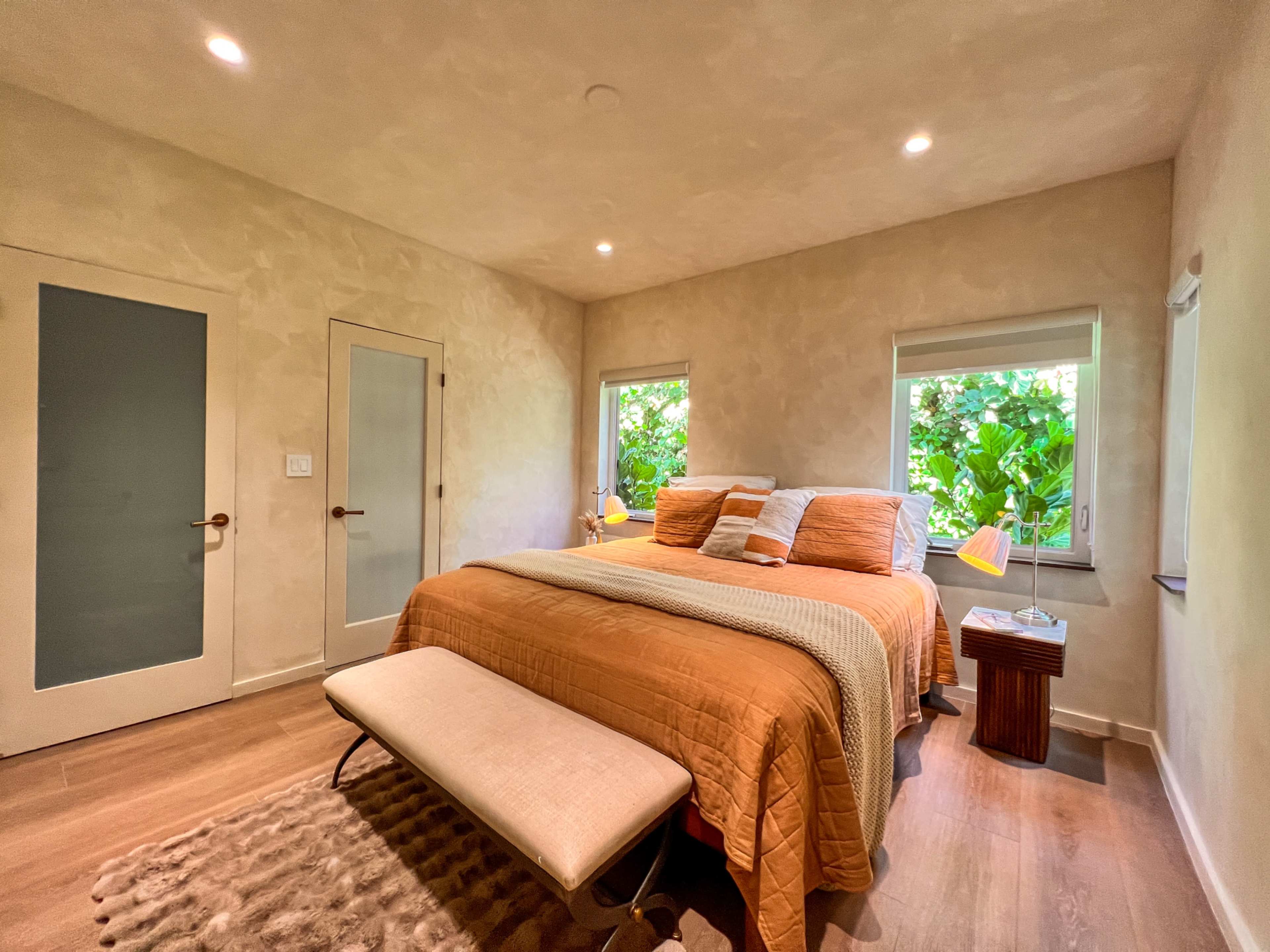 The bedroom features a large bed with orange bedding, a bench at the foot, and two windows surrounded by greenery.