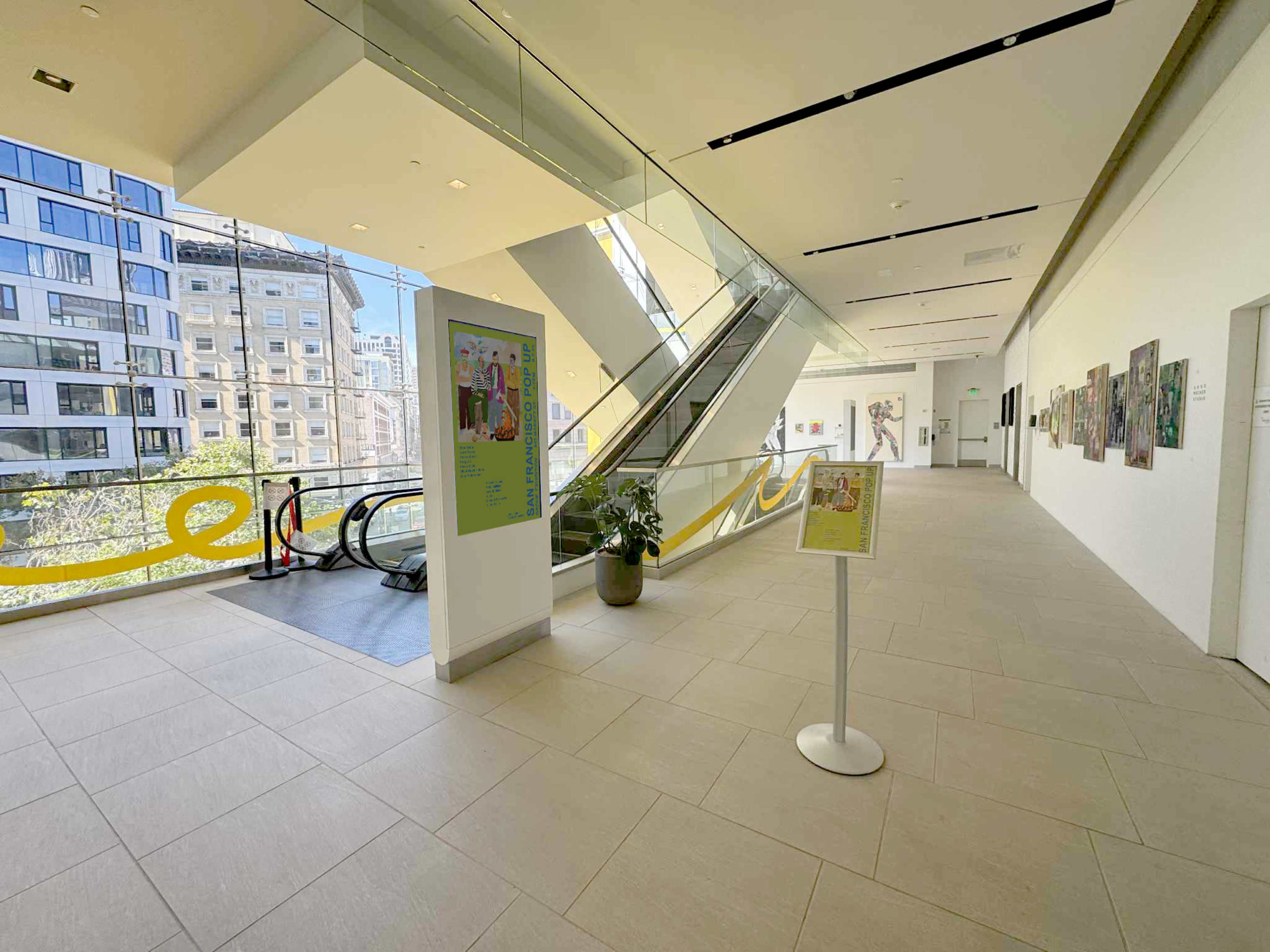 The image shows a brightly lit, modern interior of a building featuring a staircase and escalator, along with artwork displayed on the walls.