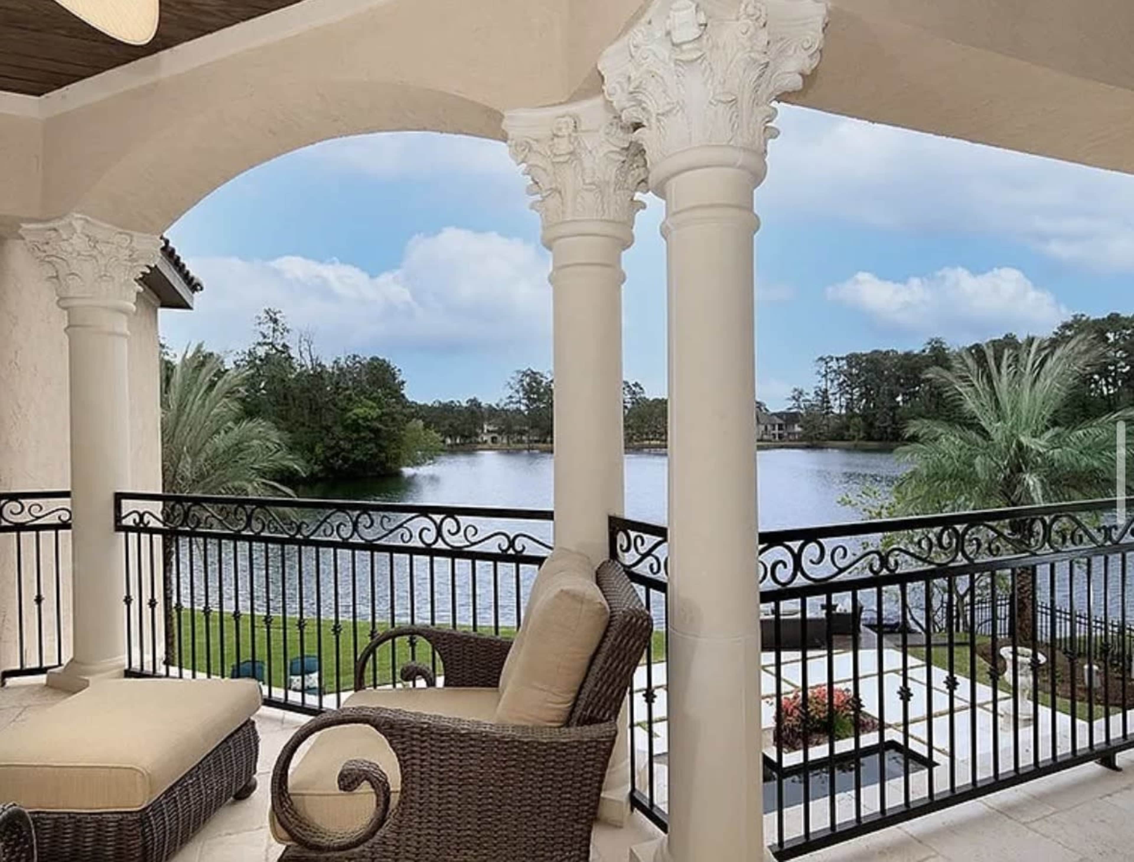 The image shows a balcony with decorative columns overlooking a tranquil body of water surrounded by trees and landscaping.