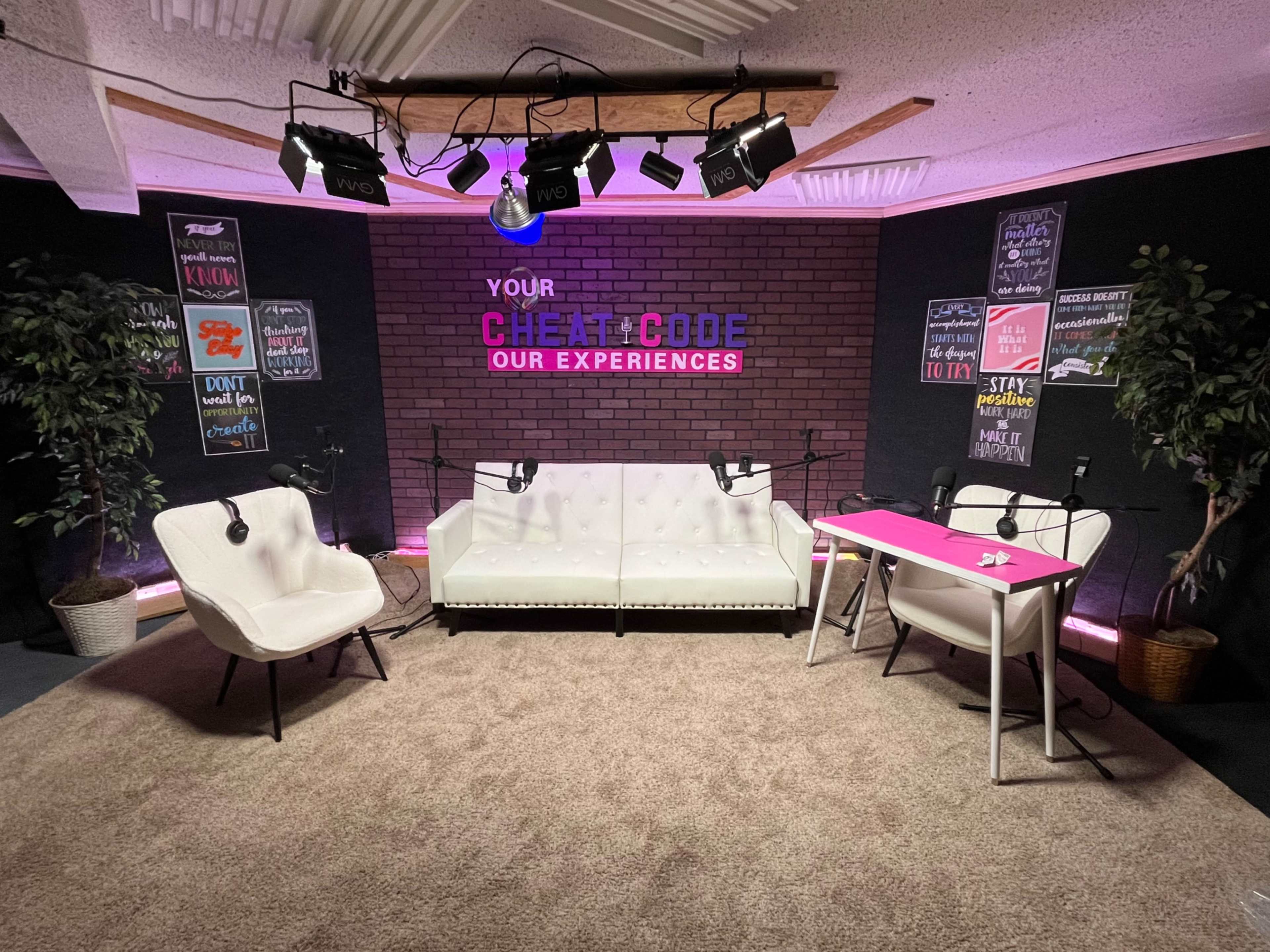 A podcast recording studio features a white couch, two chairs, and a table, accented by a brick wall and colorful signs, illuminated by overhead lights.