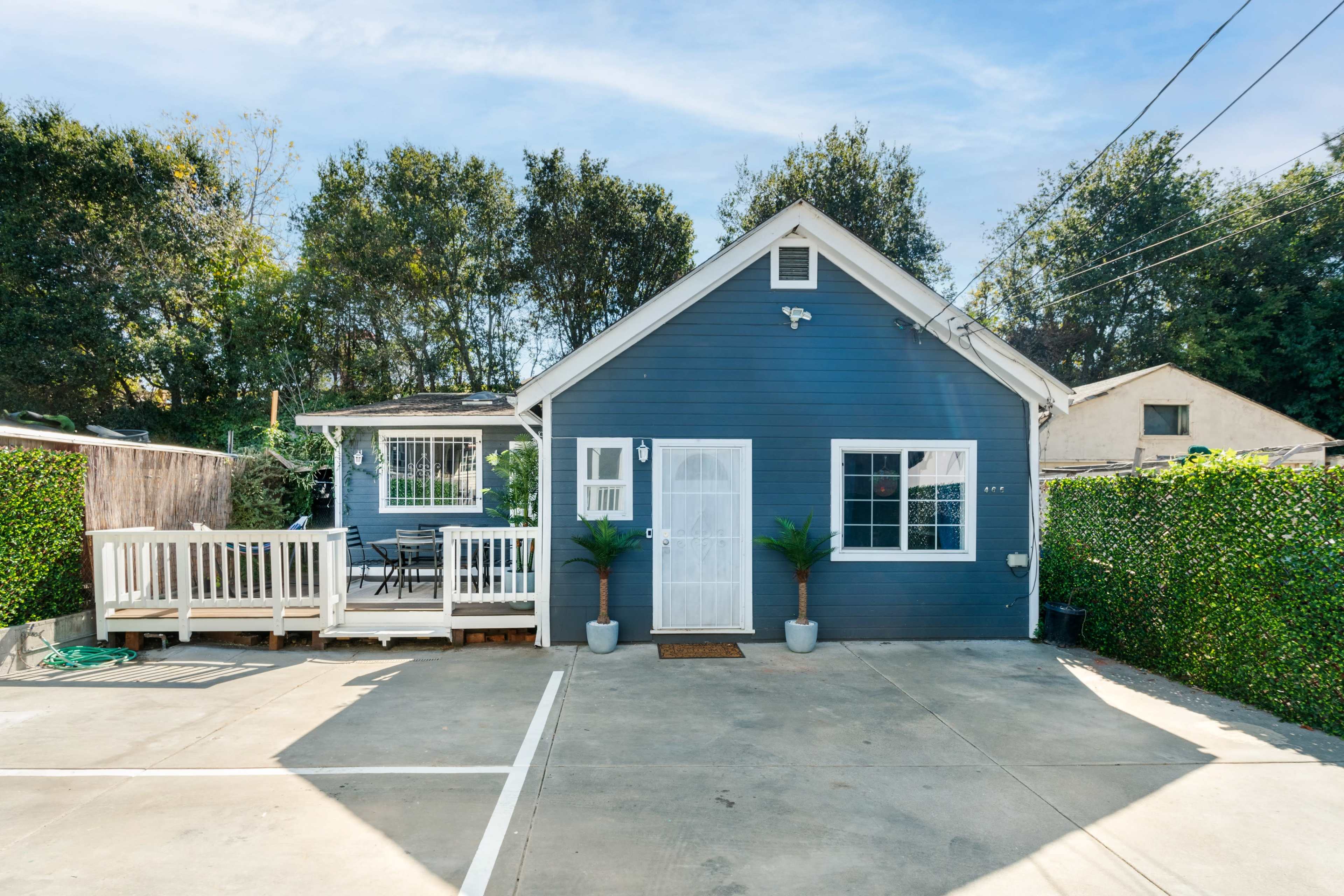 A small blue house with a white door and porch is situated next to a patio area and is surrounded by greenery.