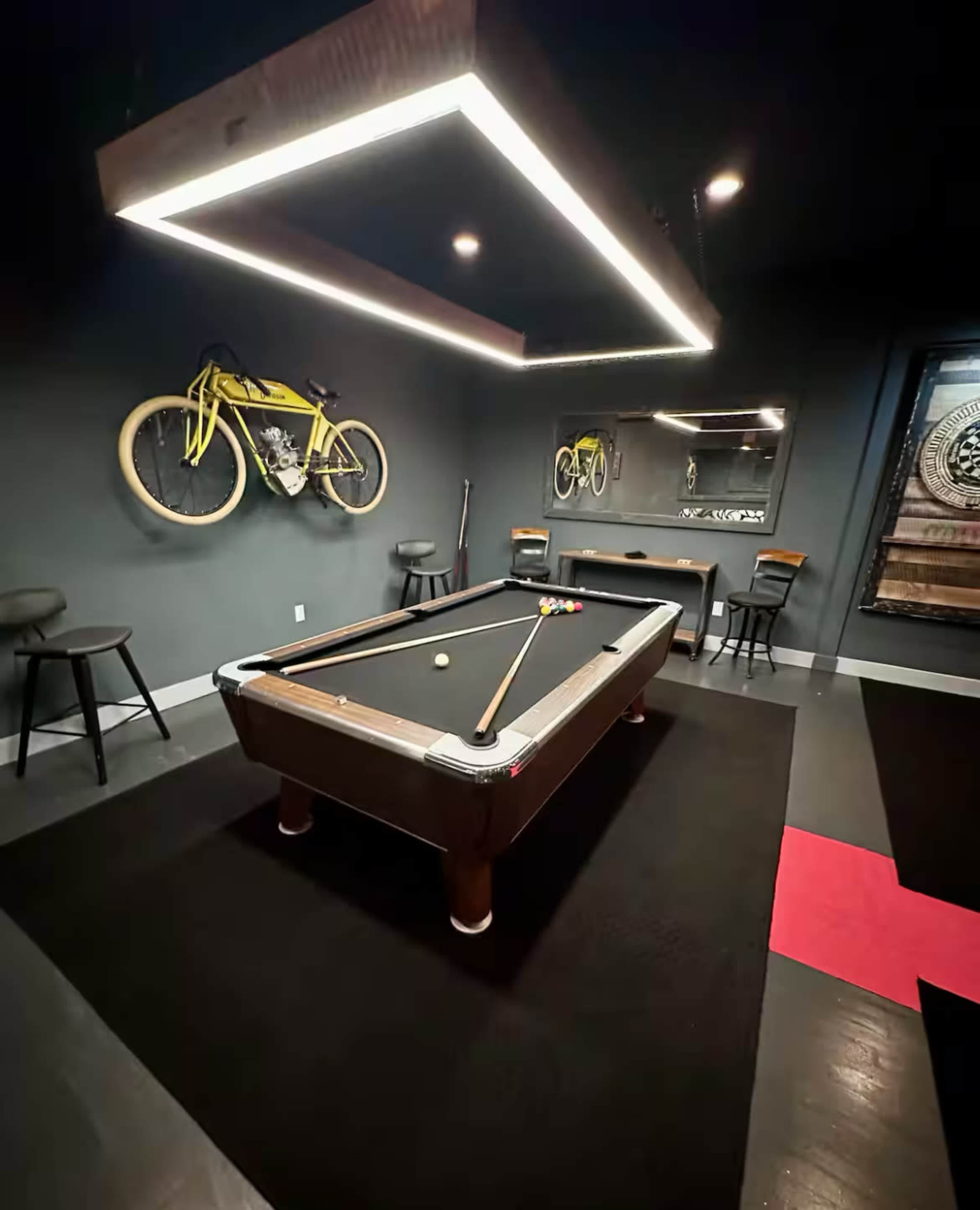 The image shows a game room featuring a billiard table, bar stools, a wall-mounted bicycle, and a mirror, all illuminated by hanging LED lights.