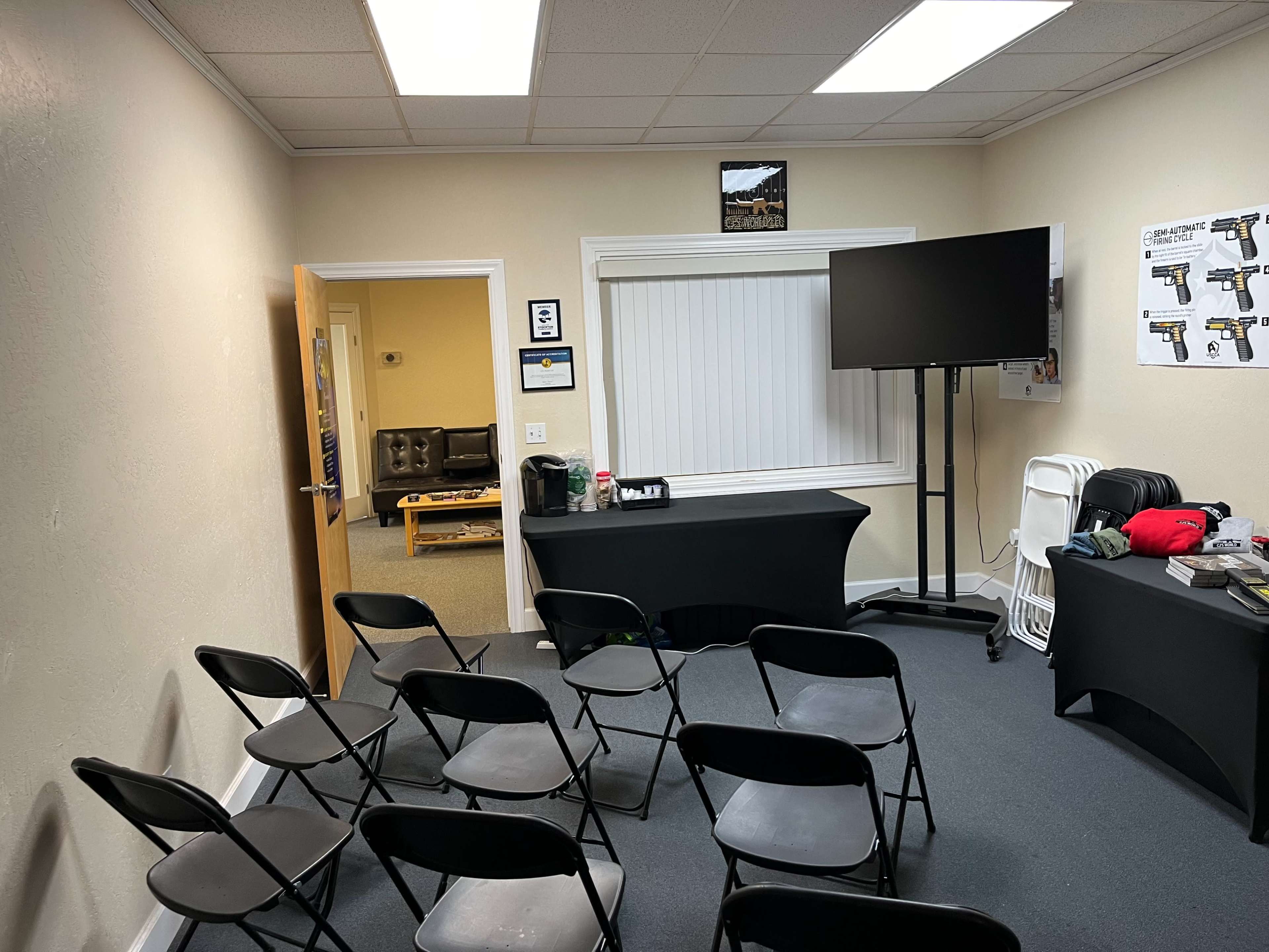 A small meeting room with black folding chairs arranged in rows, a table covered with a black cloth, and a large screen on a stand opposite a window with blinds.