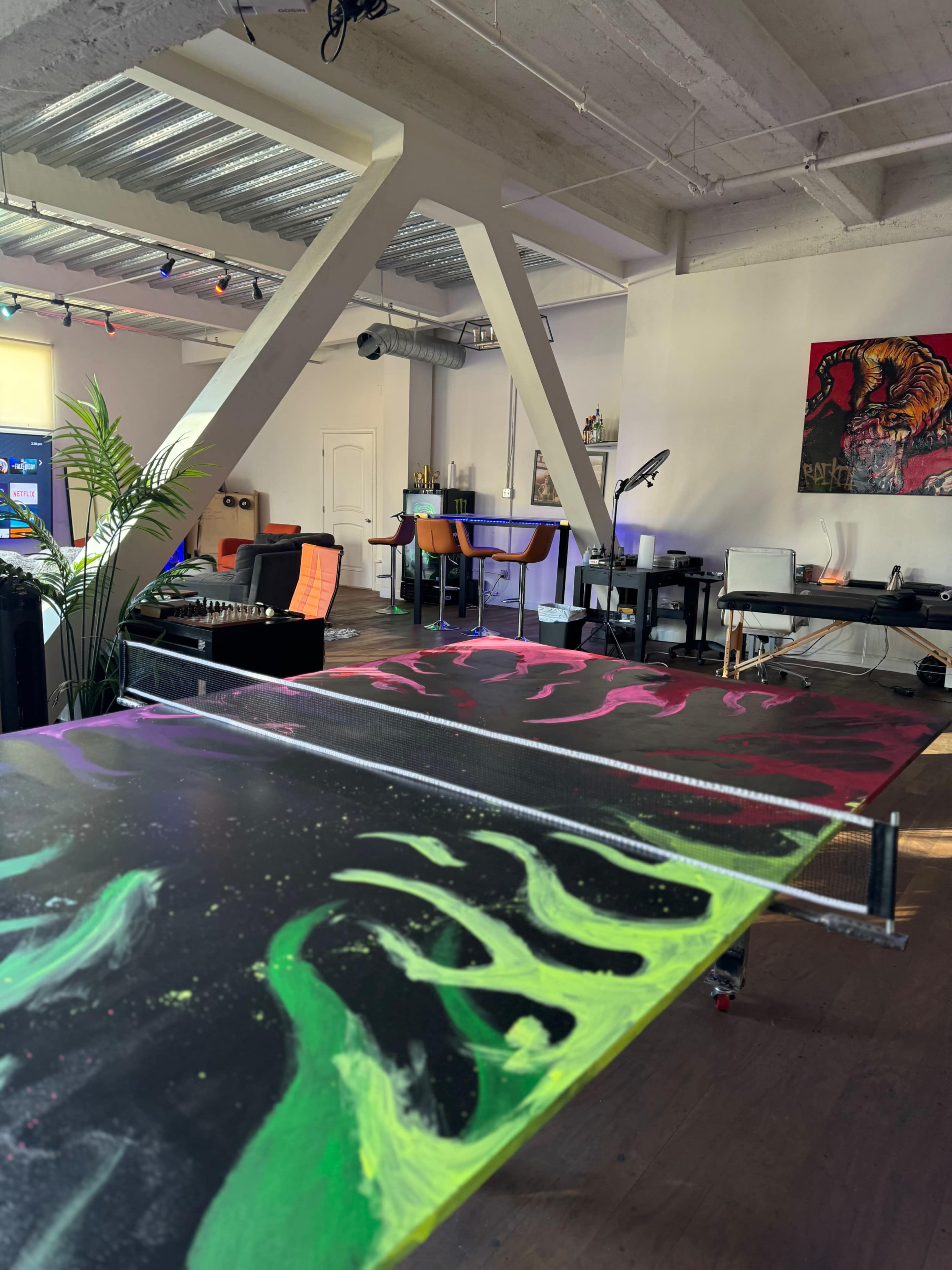 An artist's studio features a large painting on a table with vibrant colors, surrounded by various art supplies and furniture.