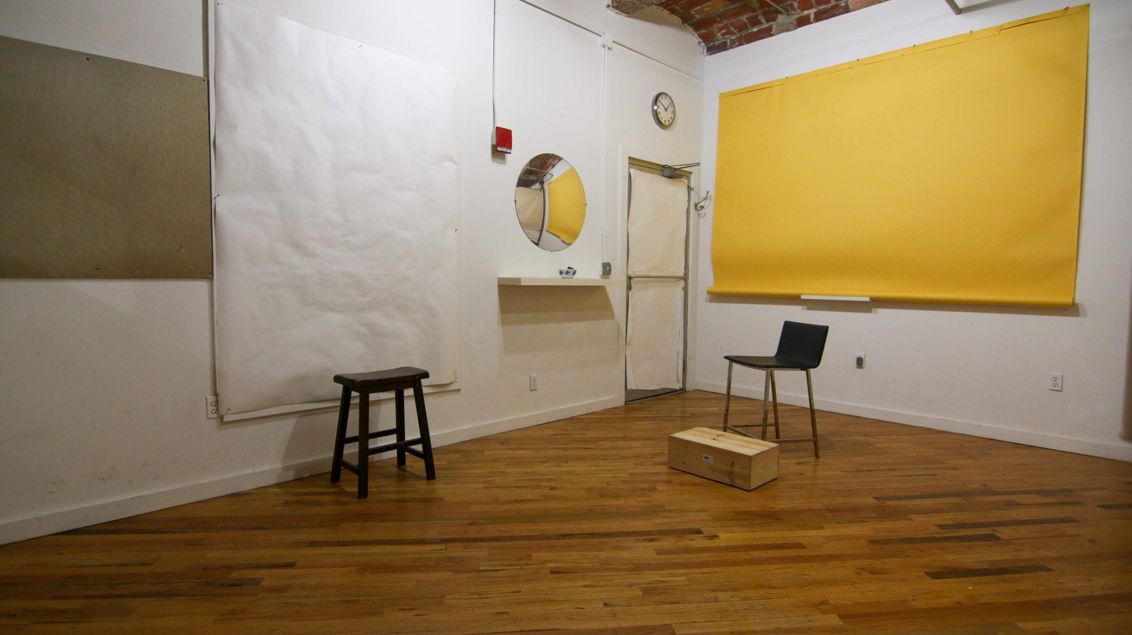 The scene depicts a sparsely furnished room with a wooden floor, featuring a stool, a black chair, a wooden box, and large sheets of white paper on the walls.