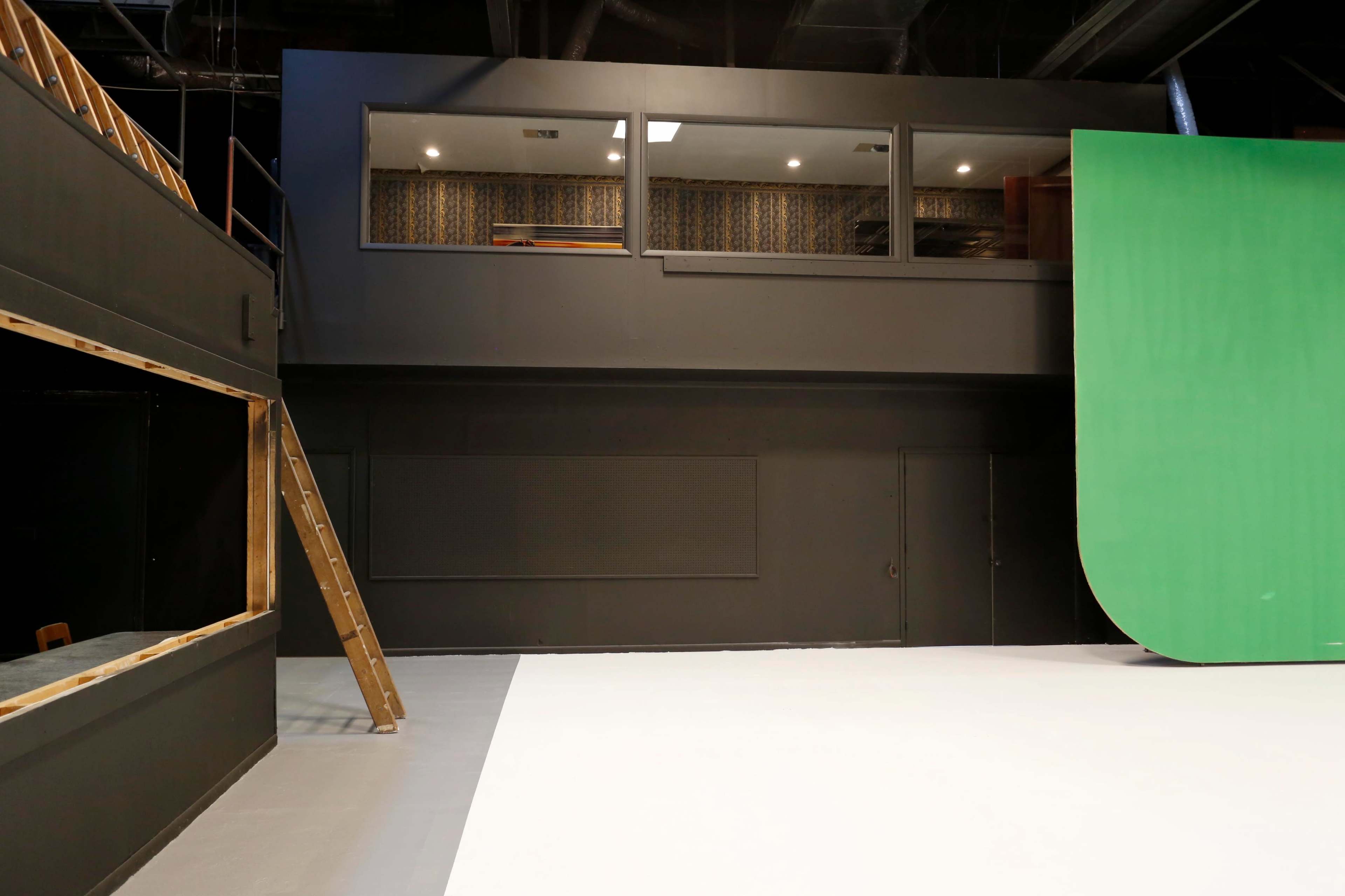A film studio interior featuring a green screen on the right and a staircase leading to a viewing area above.