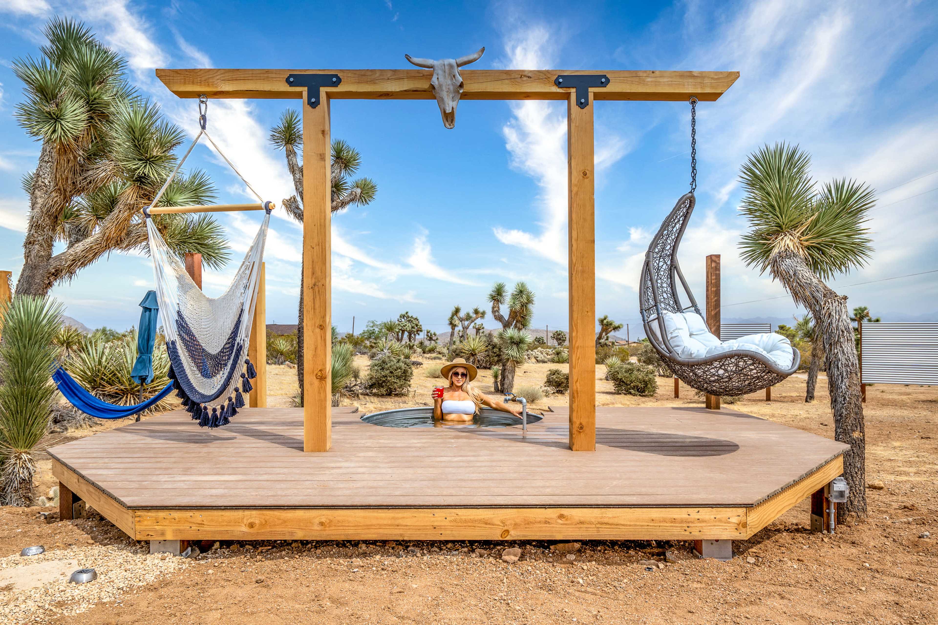 A woman relaxes in a hot tub on a wooden deck, surrounded by desert landscape and decorative swings.