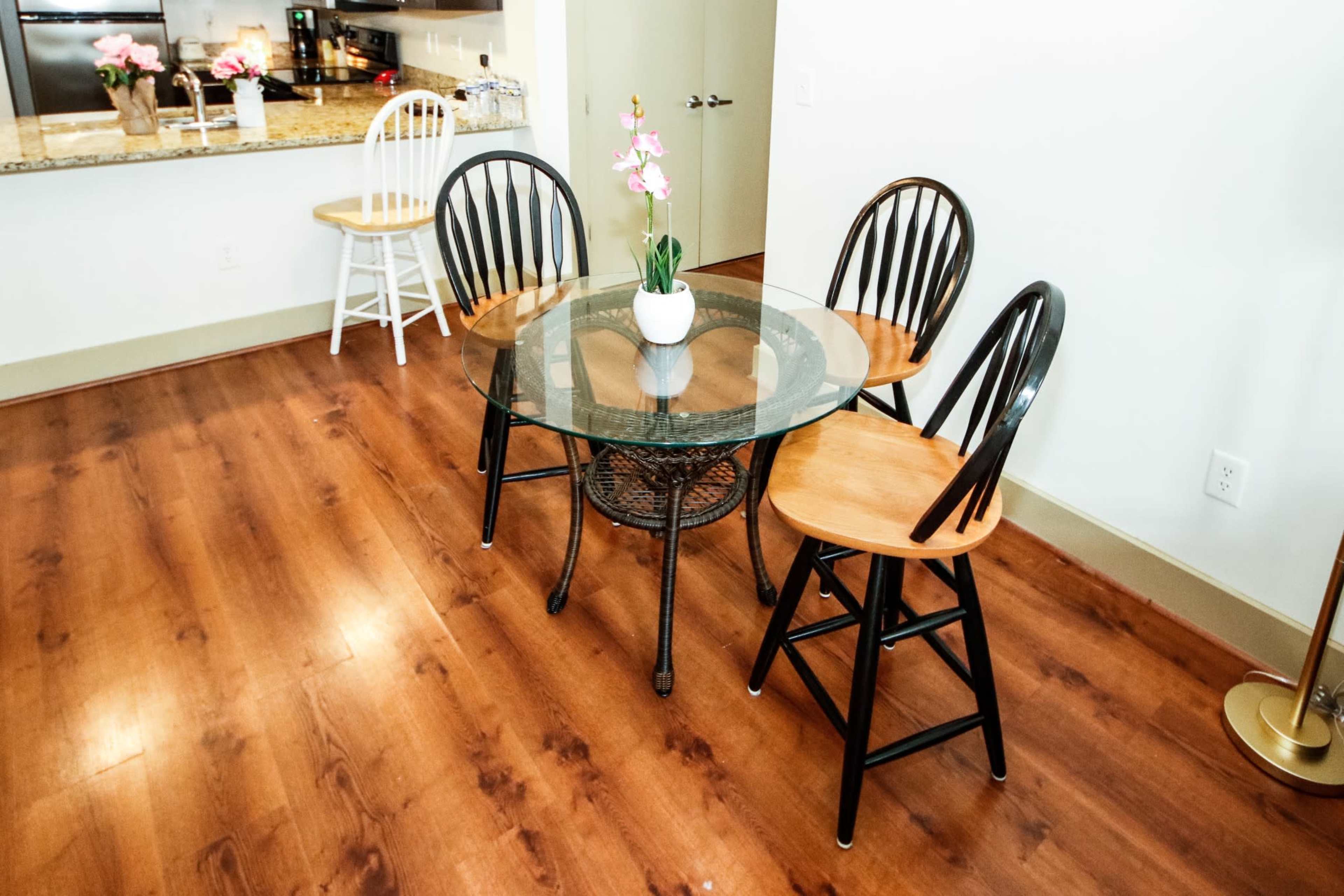 A glass-top table with four wooden chairs is set on a hardwood floor in a dining area near a kitchen.