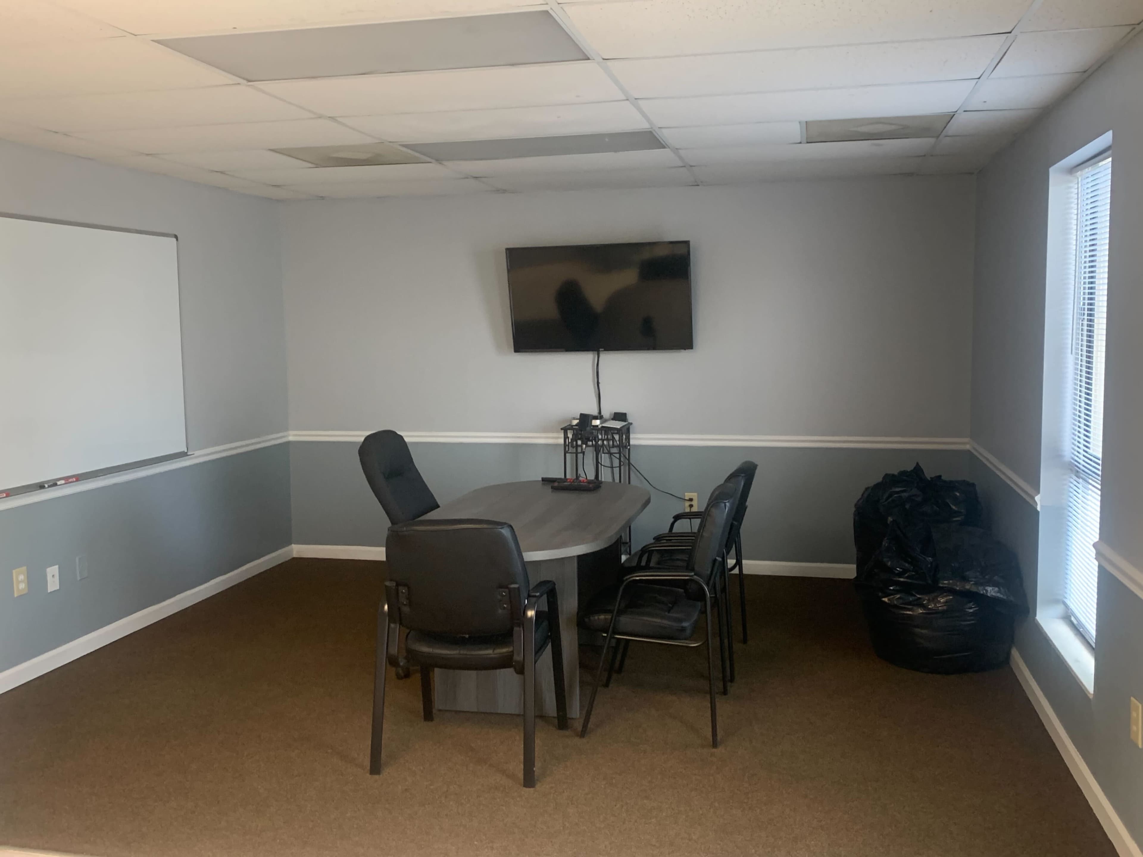 A sparsely furnished meeting room with a gray table, four black chairs, a television mounted on the wall, and a whiteboard.