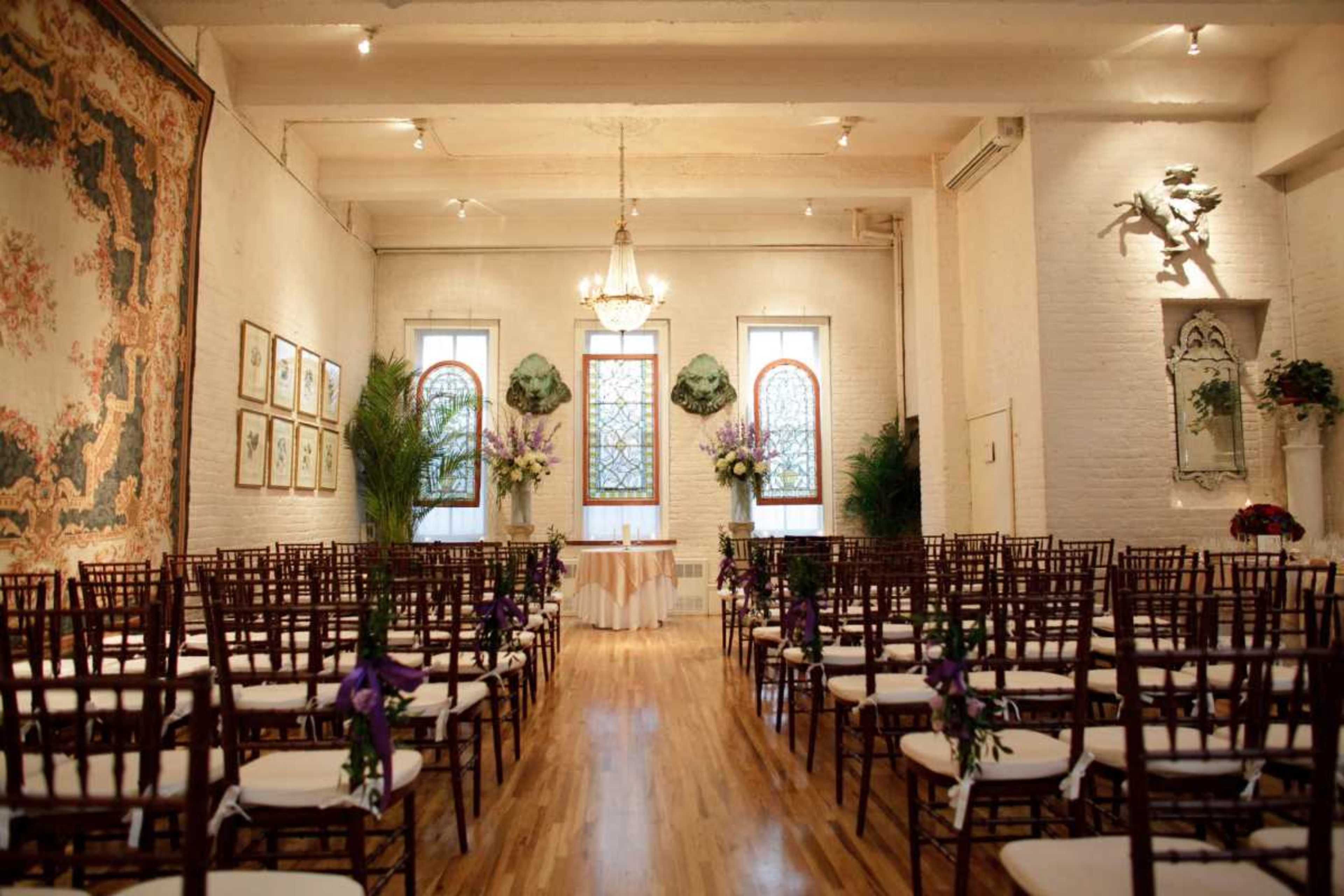 The image shows a decorated indoor venue set up for a wedding ceremony, featuring rows of wooden chairs facing a beautifully adorned altar with floral arrangements and stained glass windows.
