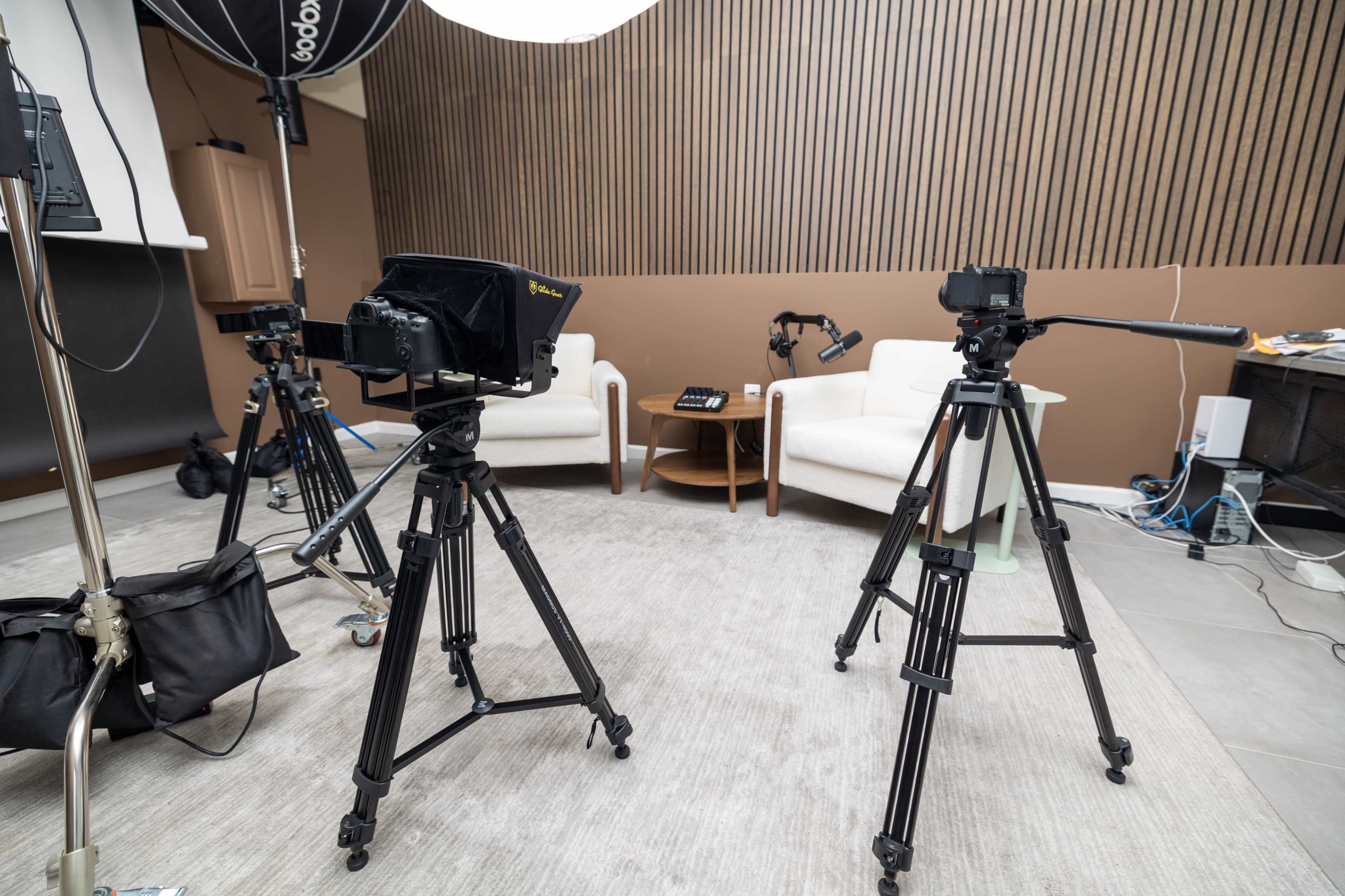 The image shows a photography setup featuring two tripods with cameras directed at an empty seating area that includes two chairs and a small table, with additional lighting equipment visible.