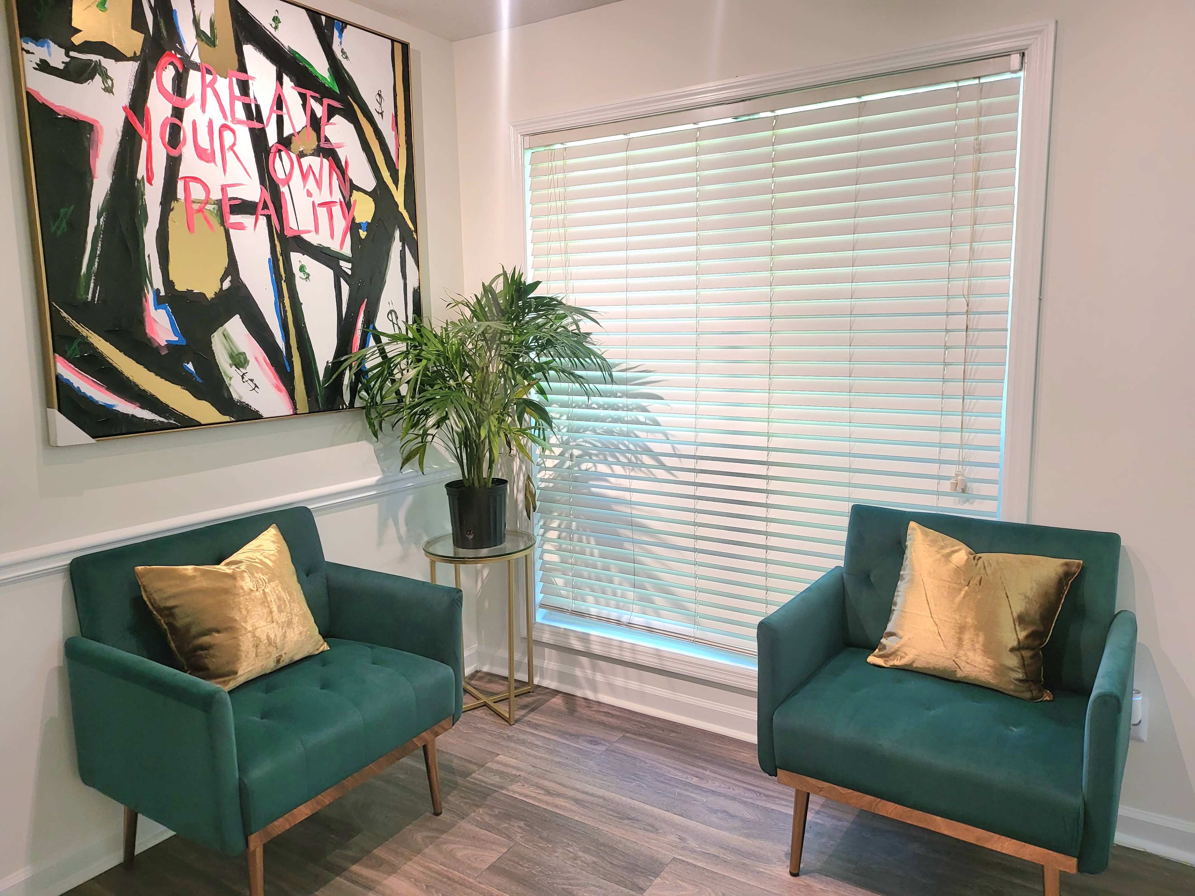 A bright room featuring two green chairs with gold cushions, a small round table with a plant, and a large painting on the wall with the words "CREATE YOUR OWN REALITY."
