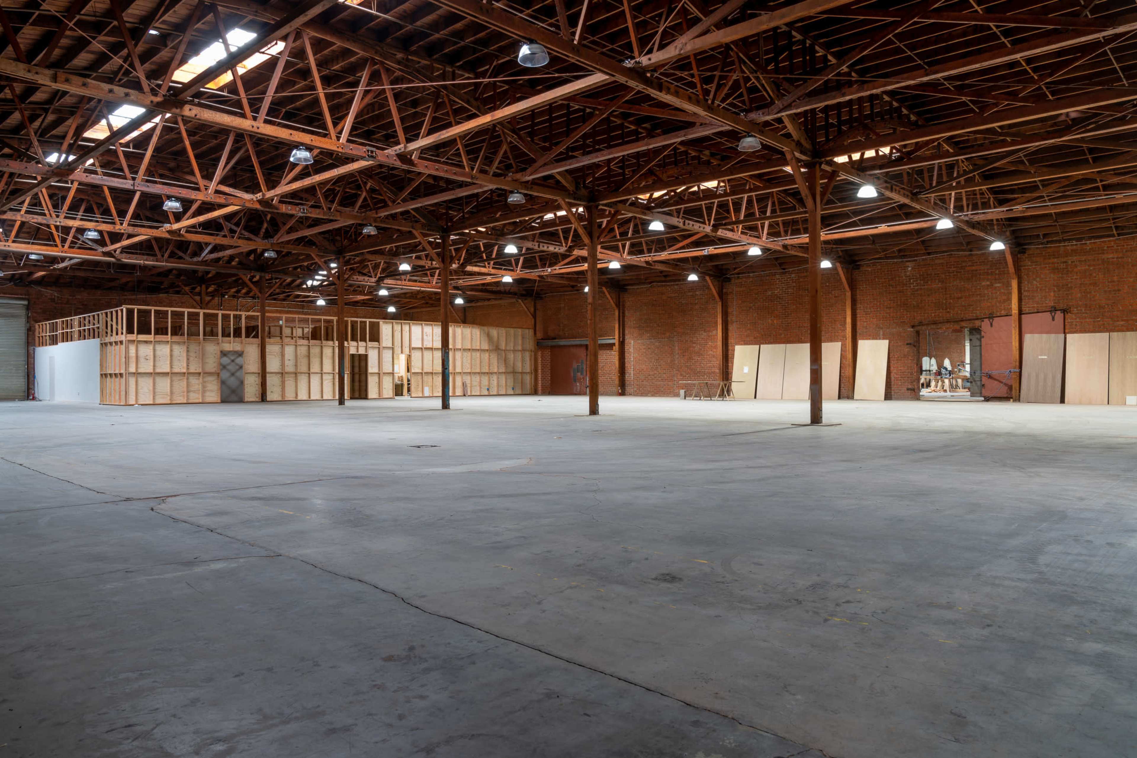 An empty warehouse with exposed brick walls and wooden beams, featuring concrete flooring and partitions made of wood in the background.