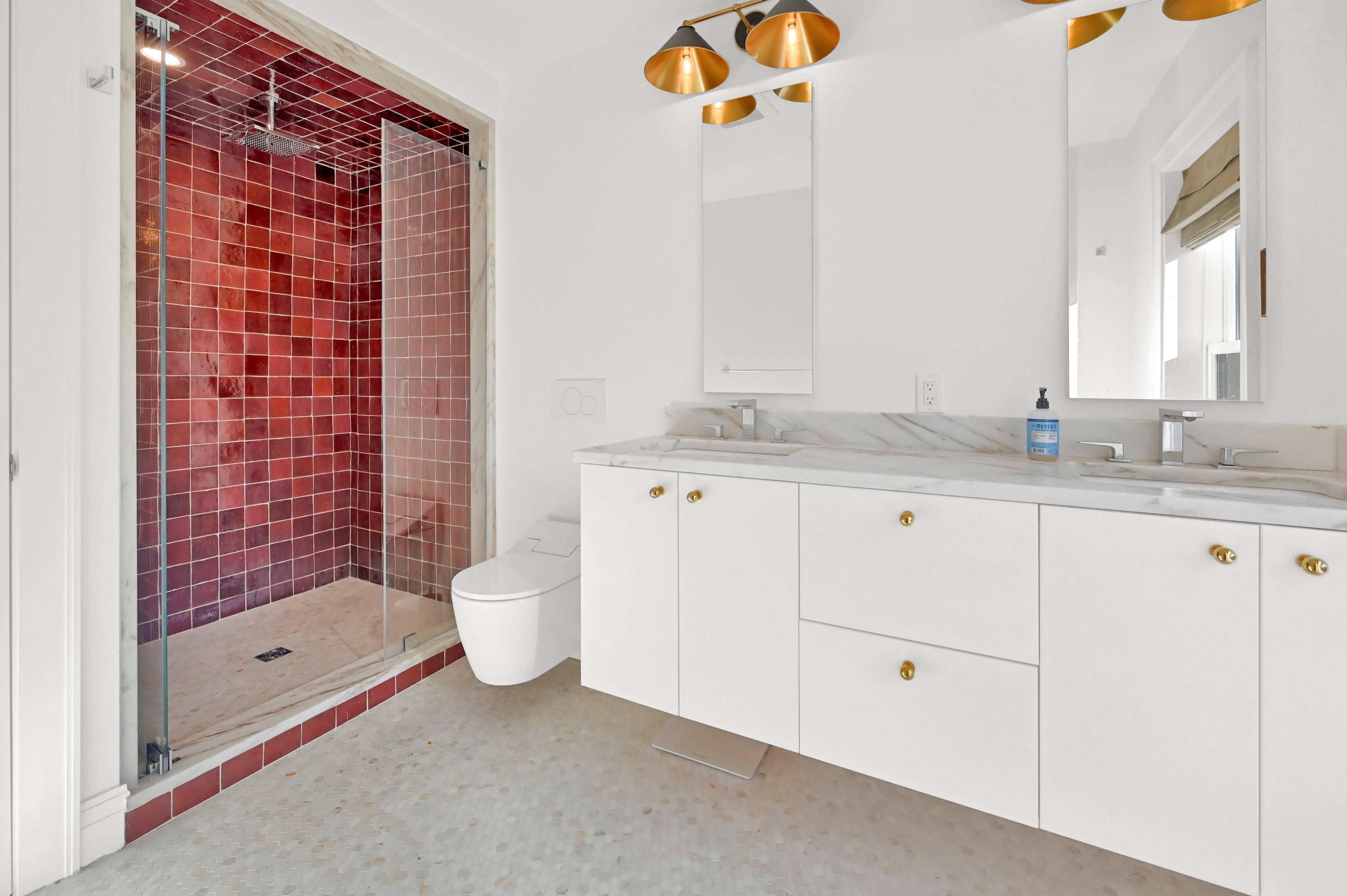 A modern bathroom featuring a shower with red tile, a white vanity with gold handles, and a large mirror.