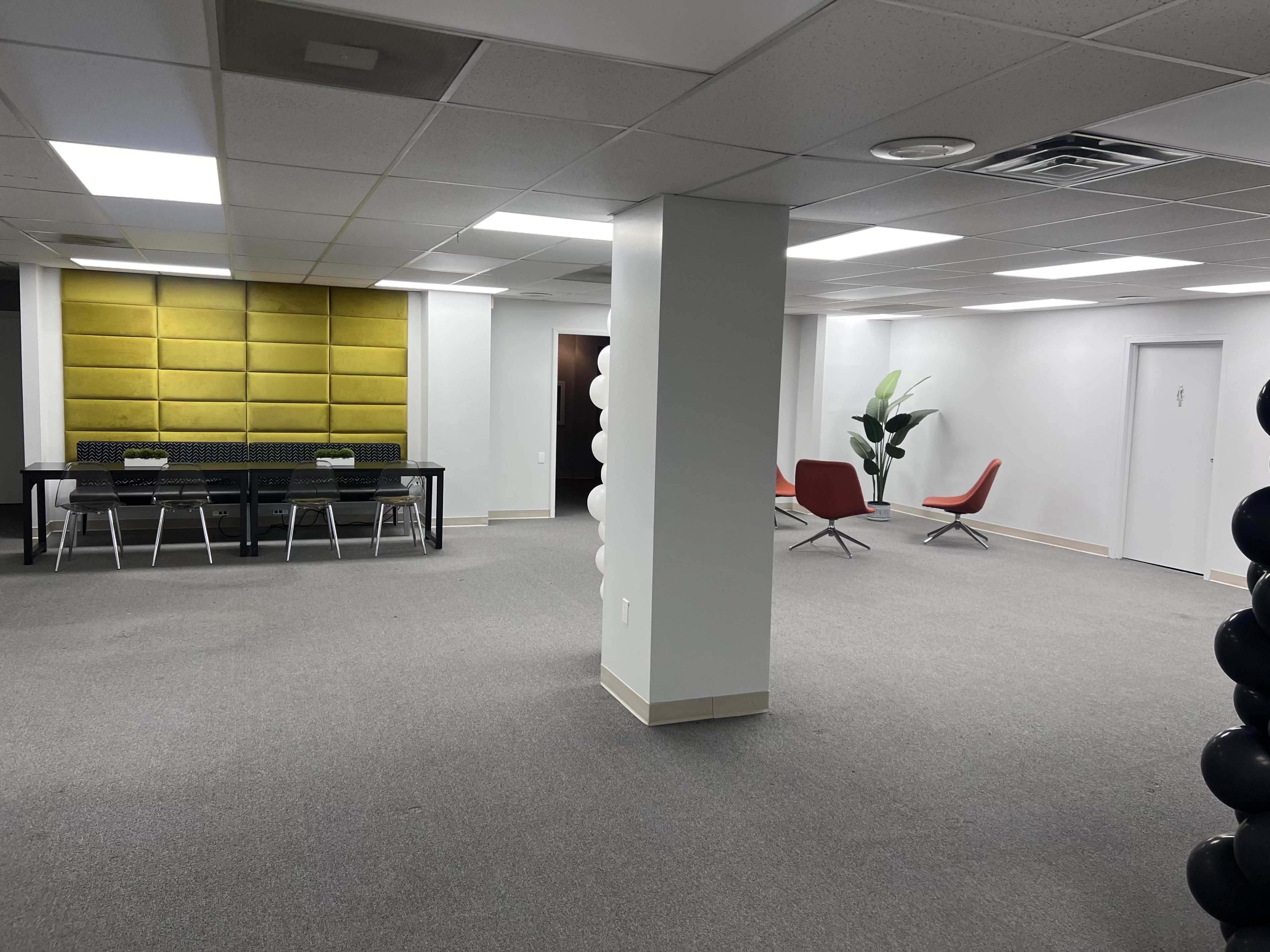 The image shows an empty office space featuring a seating area with red chairs, a large table with chairs, and a bright yellow accent wall.