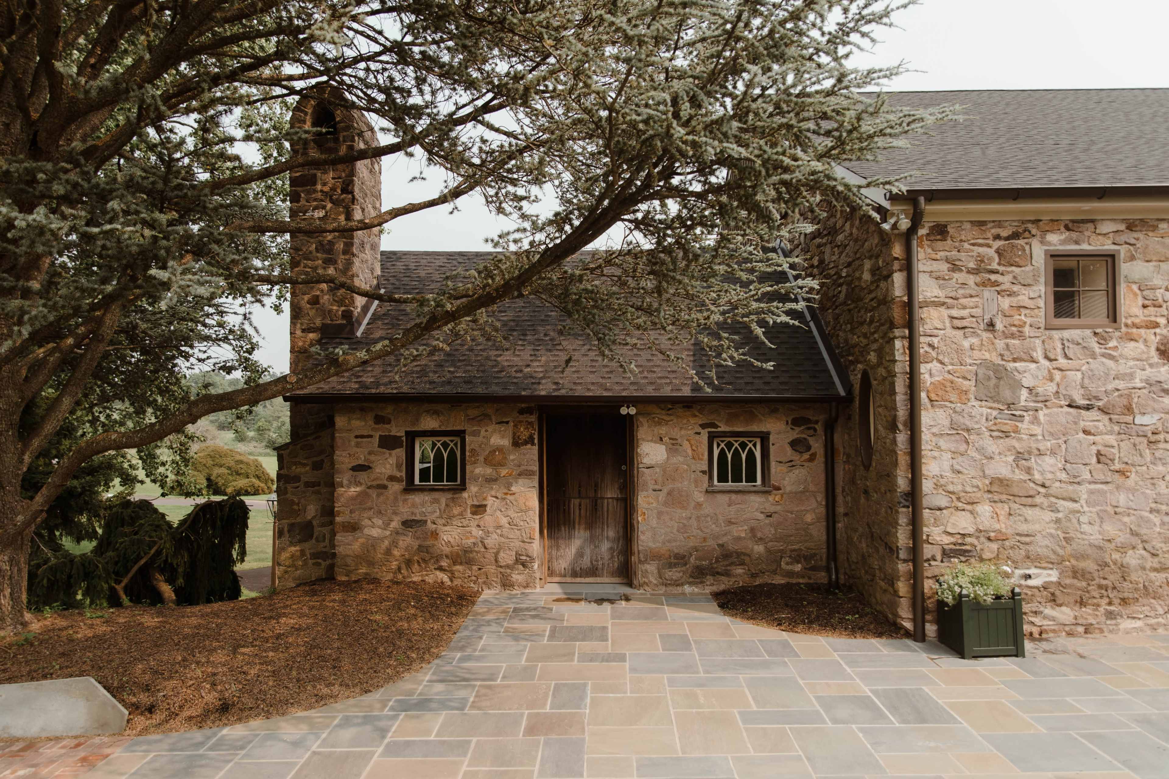 The image shows a rustic stone building with a wooden door, flanked by two small windows, set in a landscaped area with a paved pathway.