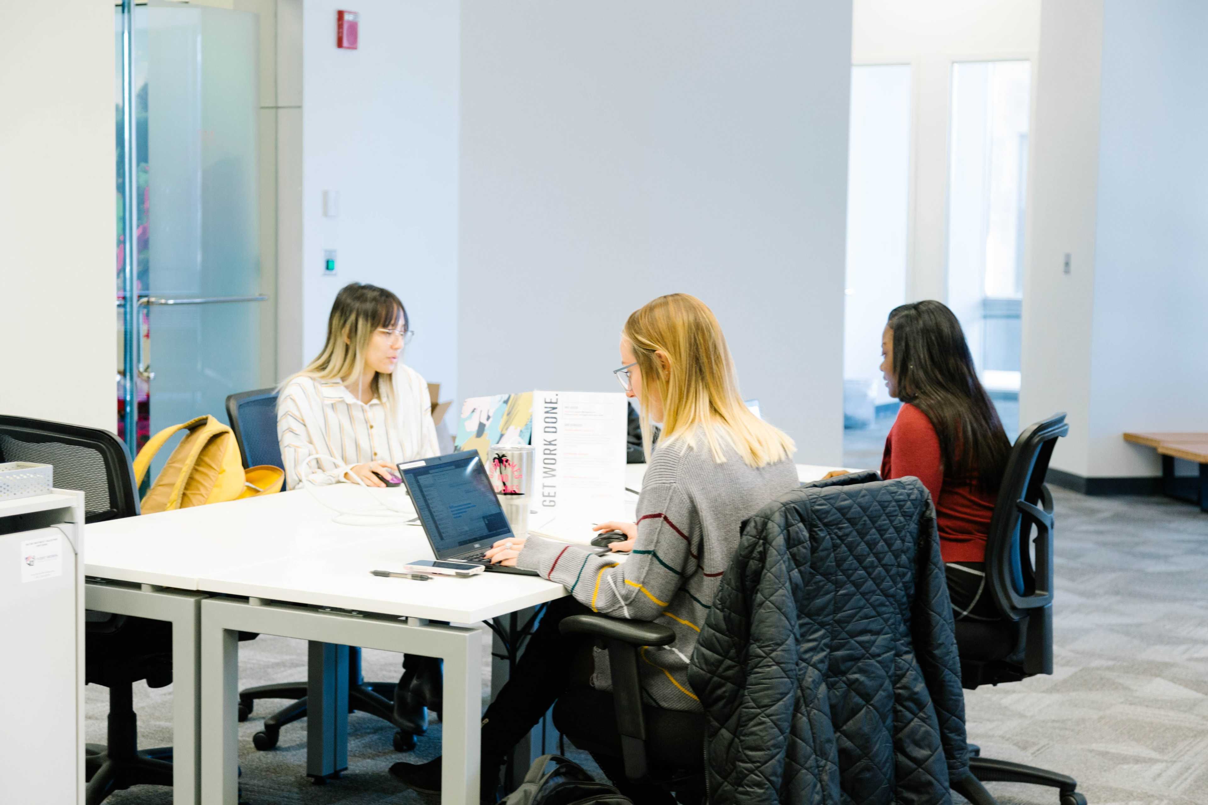 Three individuals work at a shared office space with laptops and a sign that says "I GET WORK DONE."