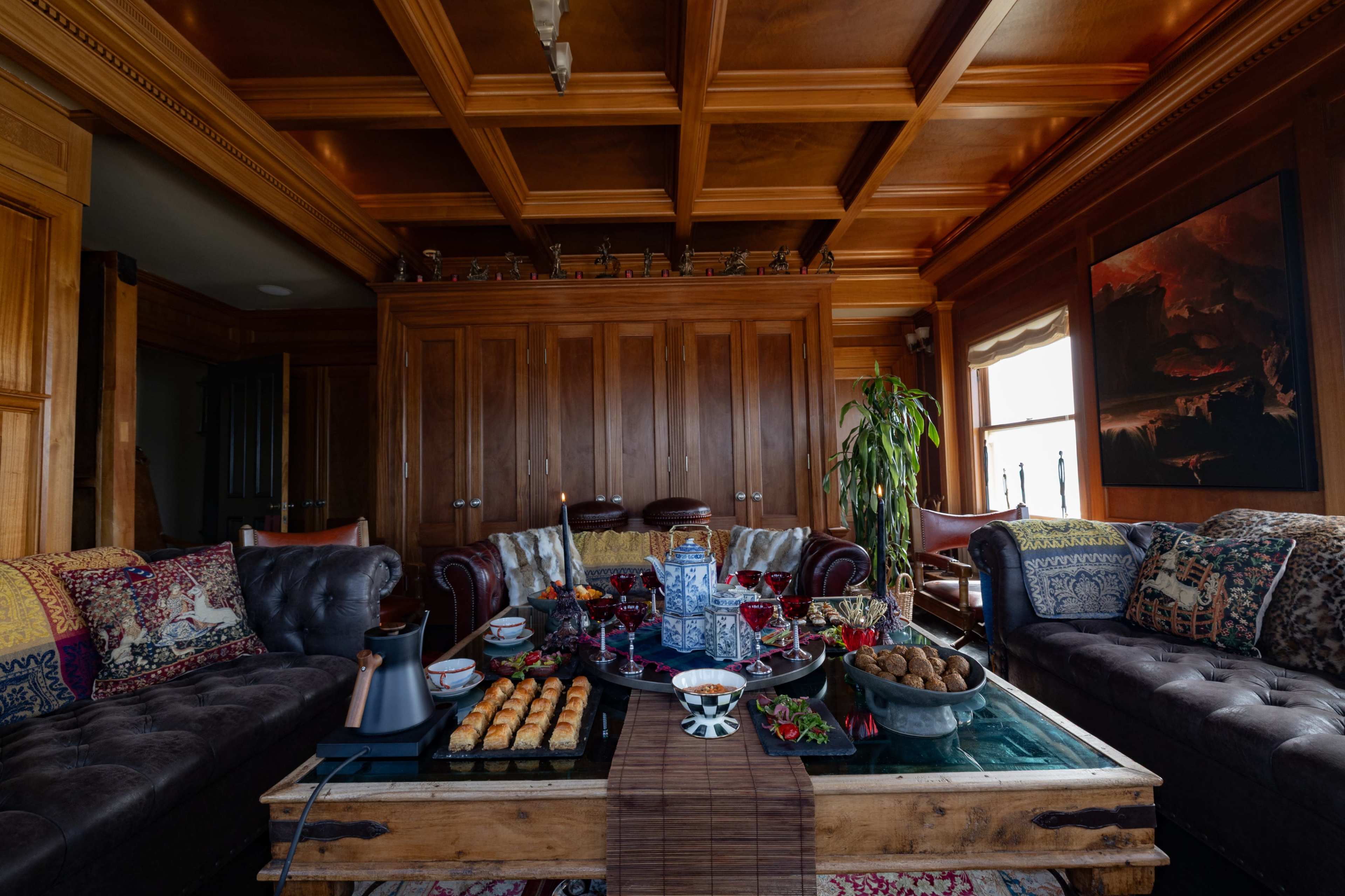 A cozy living room features two dark leather sofas facing a coffee table adorned with various dishes, drinks, and decorative items beneath a wooden coffered ceiling.