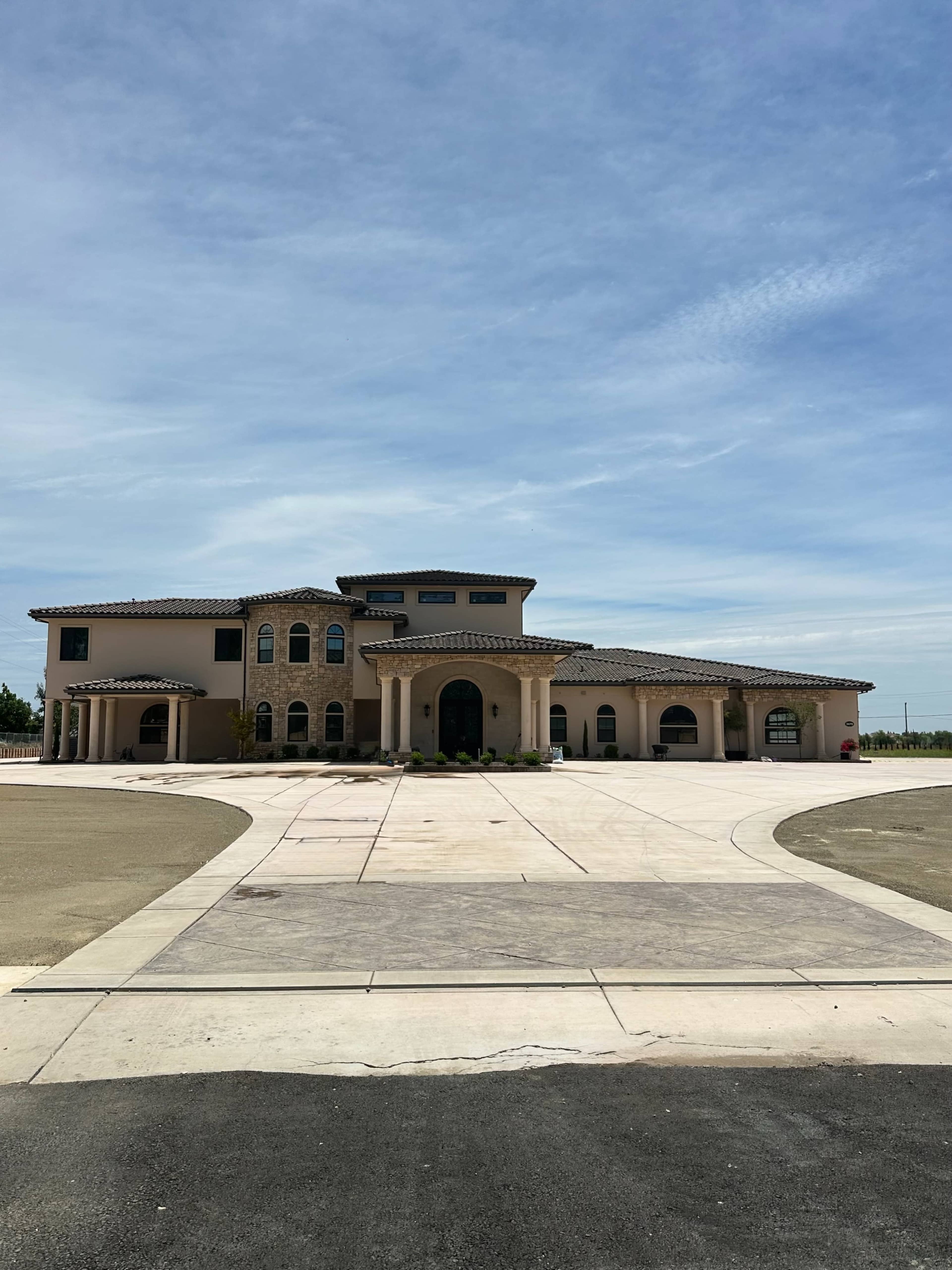 The image shows a large, modern Mediterranean-style house with a curved driveway and a clear blue sky.