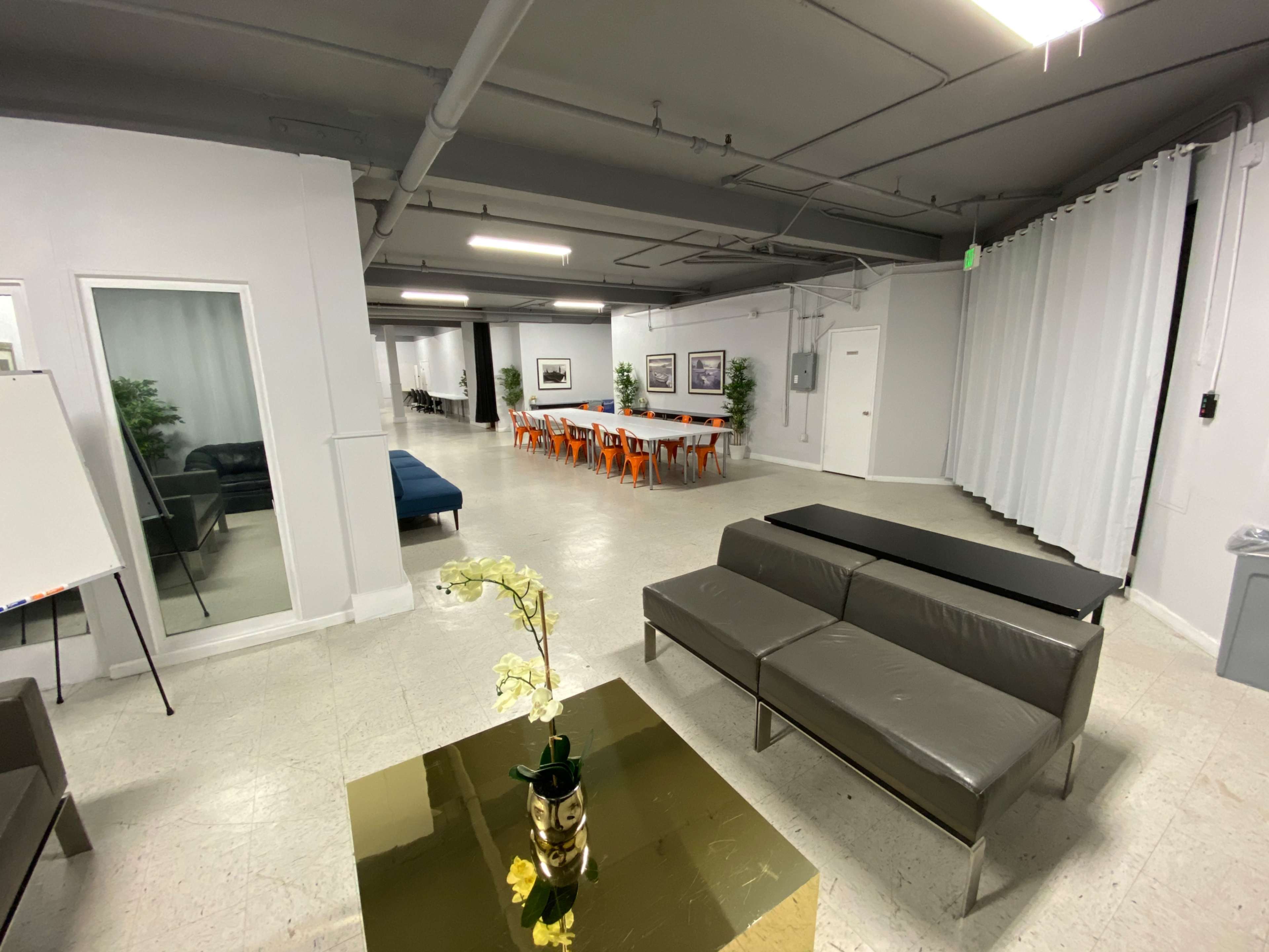 A spacious, modern room featuring a seating area with a gray sofa, a coffee table with a vase, and a dining area with orange chairs and a large table.