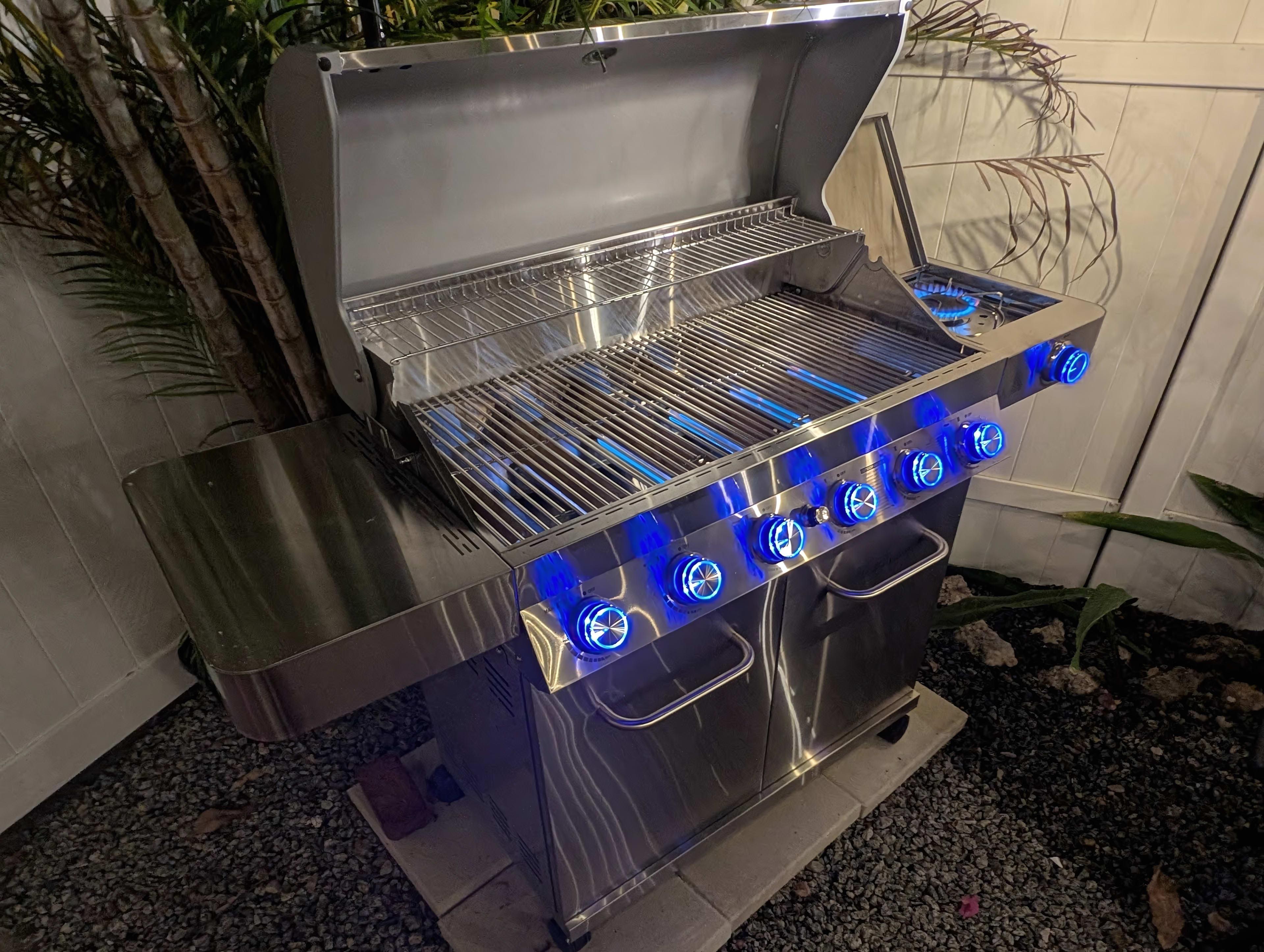 The image shows a stainless steel gas grill with five burners, illuminated control knobs, and a side shelf, positioned against a backdrop of greenery and a white fence.