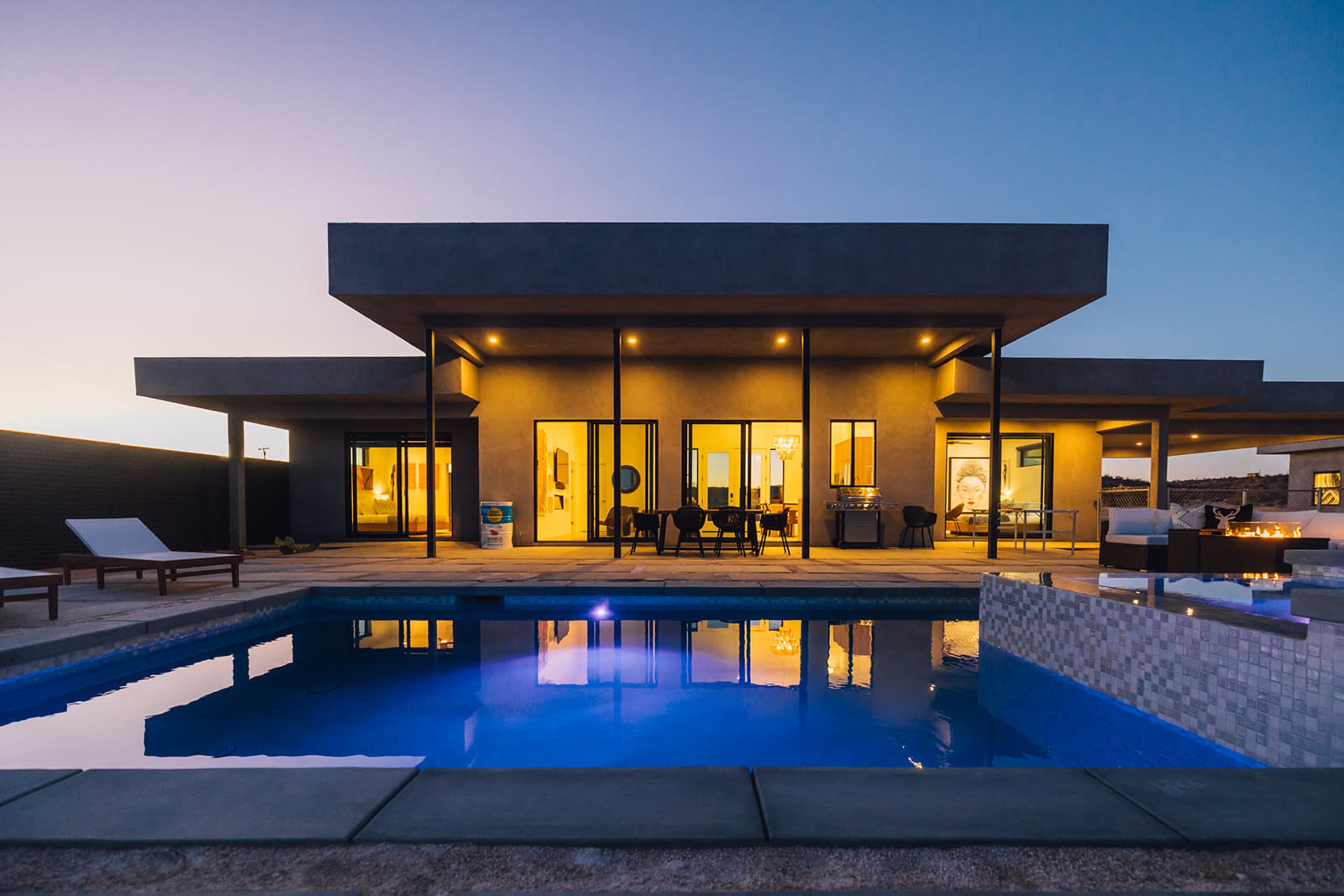 A modern house with large windows overlooks a swimming pool at dusk.