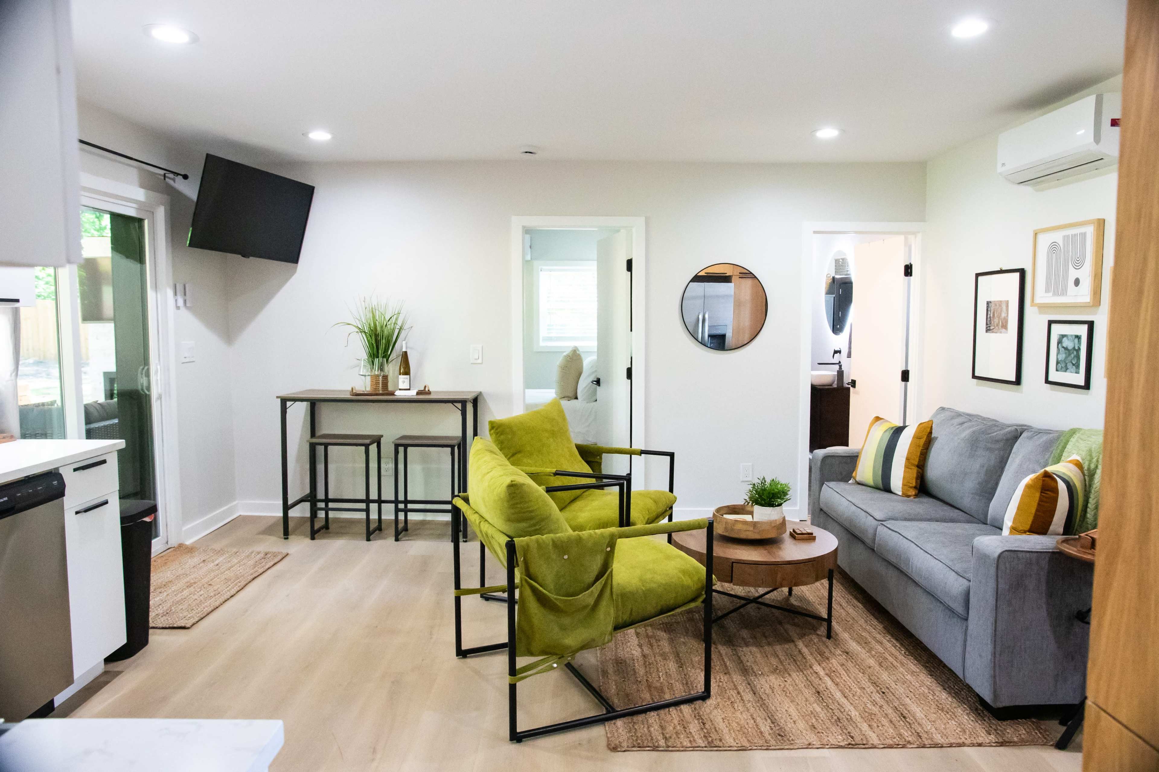 A modern living area featuring a gray sofa, two green chairs, a small dining table, and a flat-screen TV mounted on the wall.