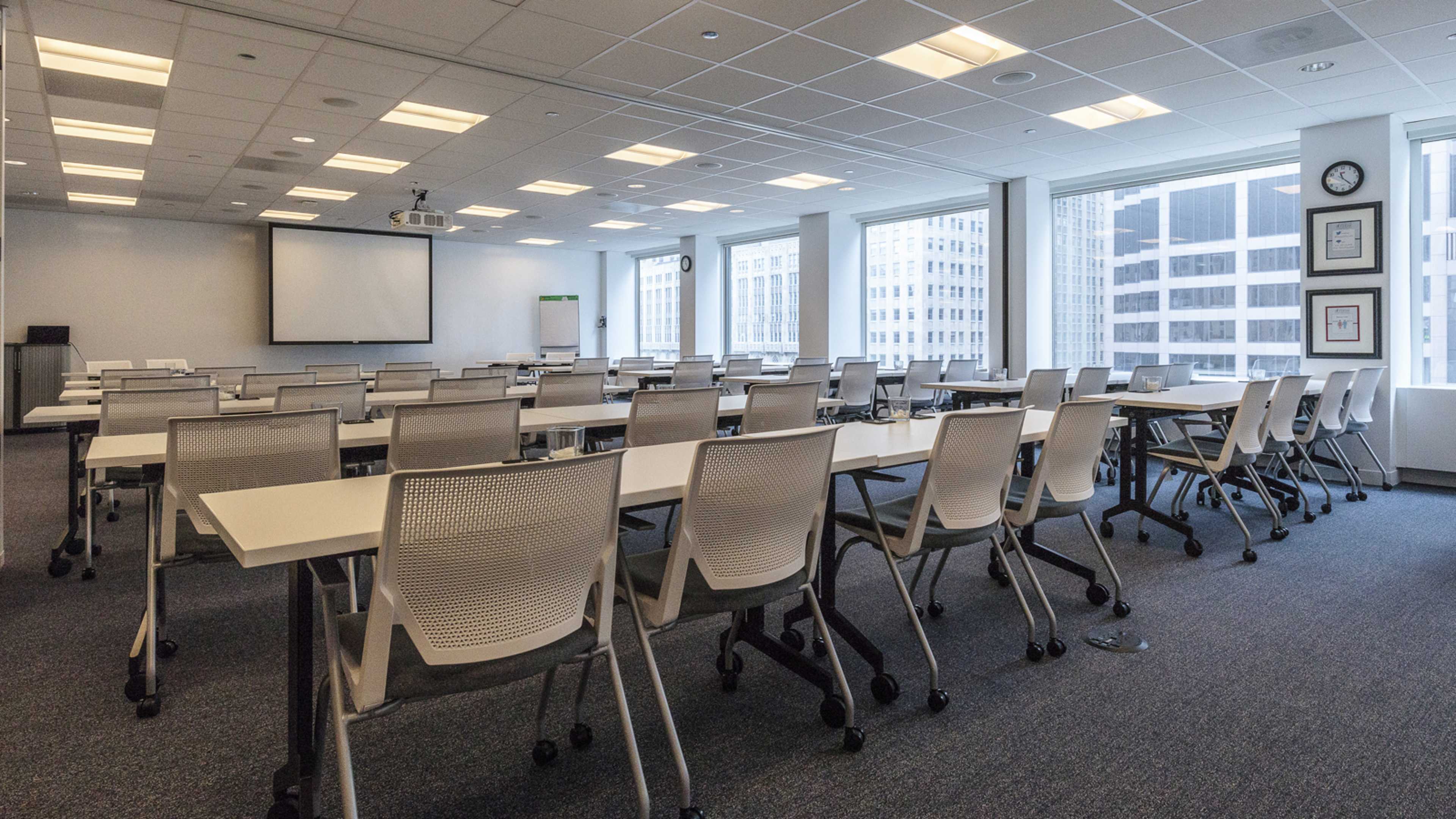 A well-lit conference room with multiple rows of tables and chairs, equipped with a projector screen at the front and large windows overlooking a cityscape.