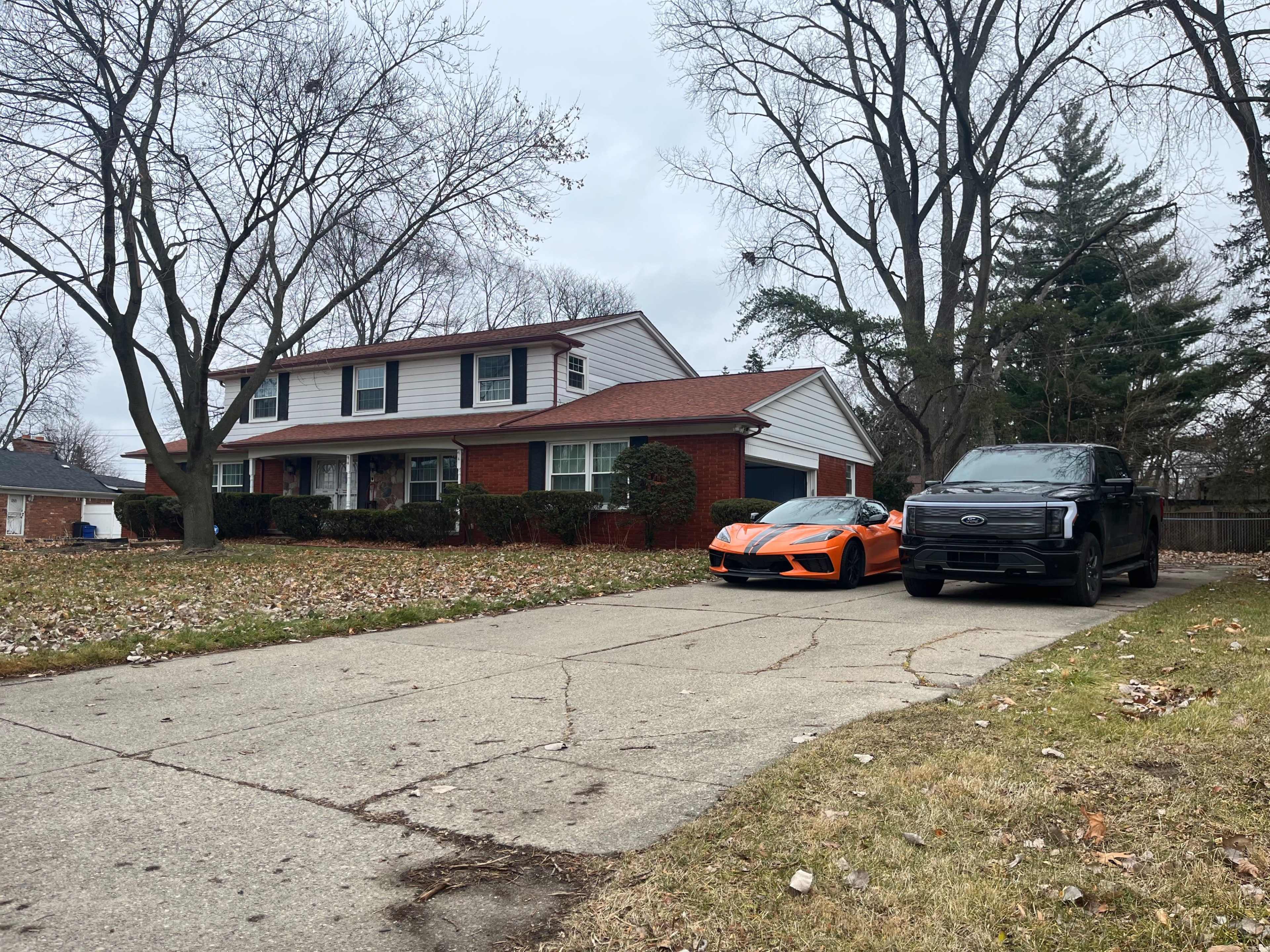 A two-story house with a red brick exterior sits next to an orange sports car and a black pickup truck on a driveway lined with uneven concrete.