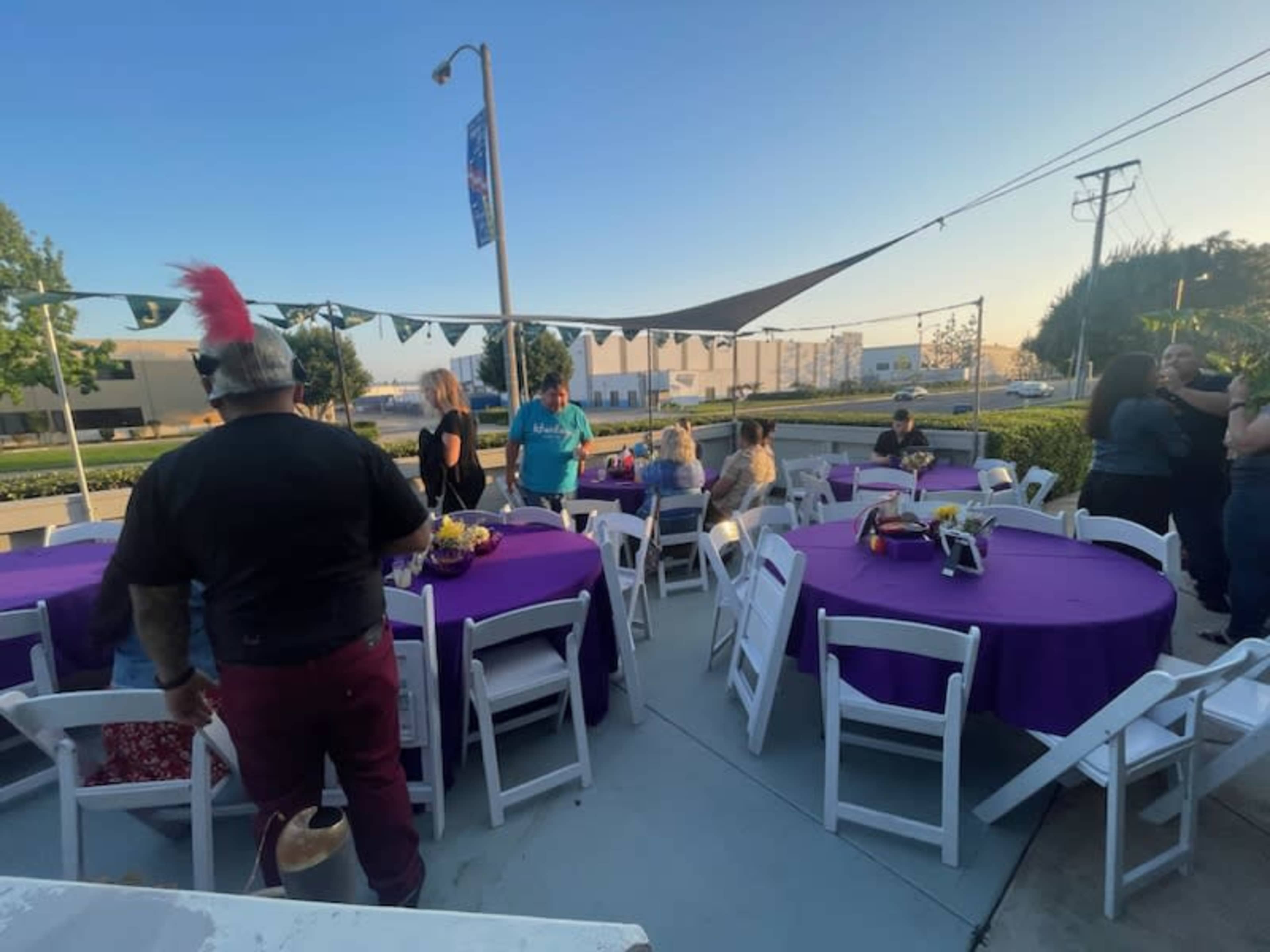 A group of people gathers around purple tablecloths at an outdoor event space with several white folding chairs.