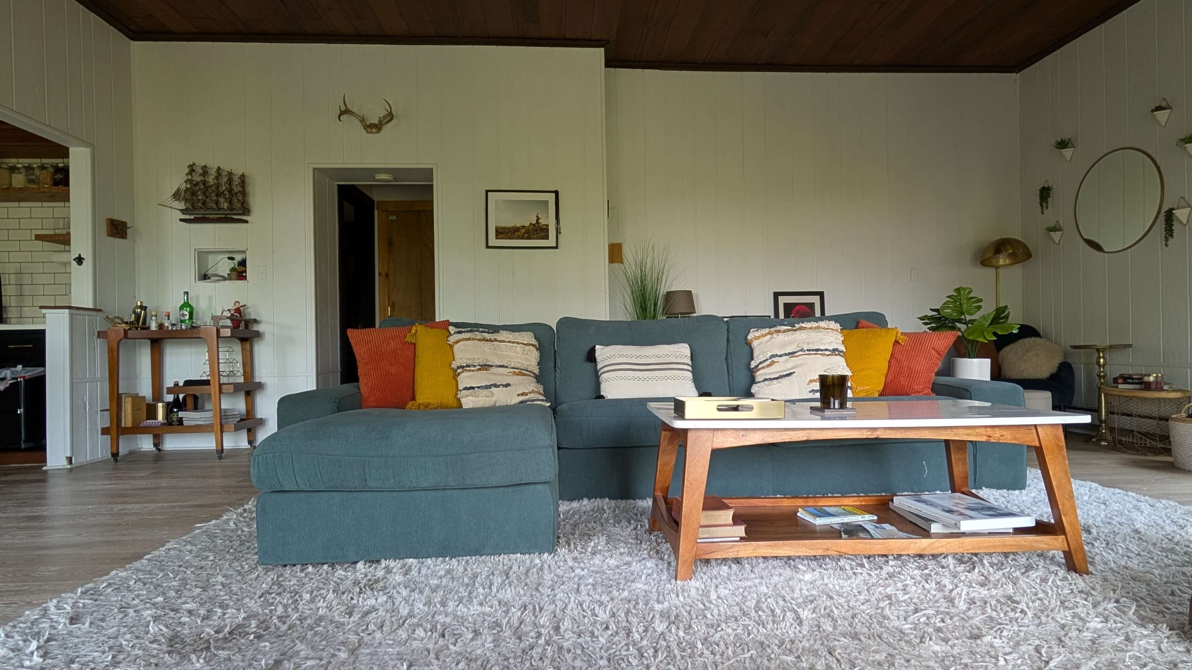 The image shows a living room featuring a large blue sectional sofa with colorful accent pillows, a wooden coffee table, and a light rug on a hardwood floor.