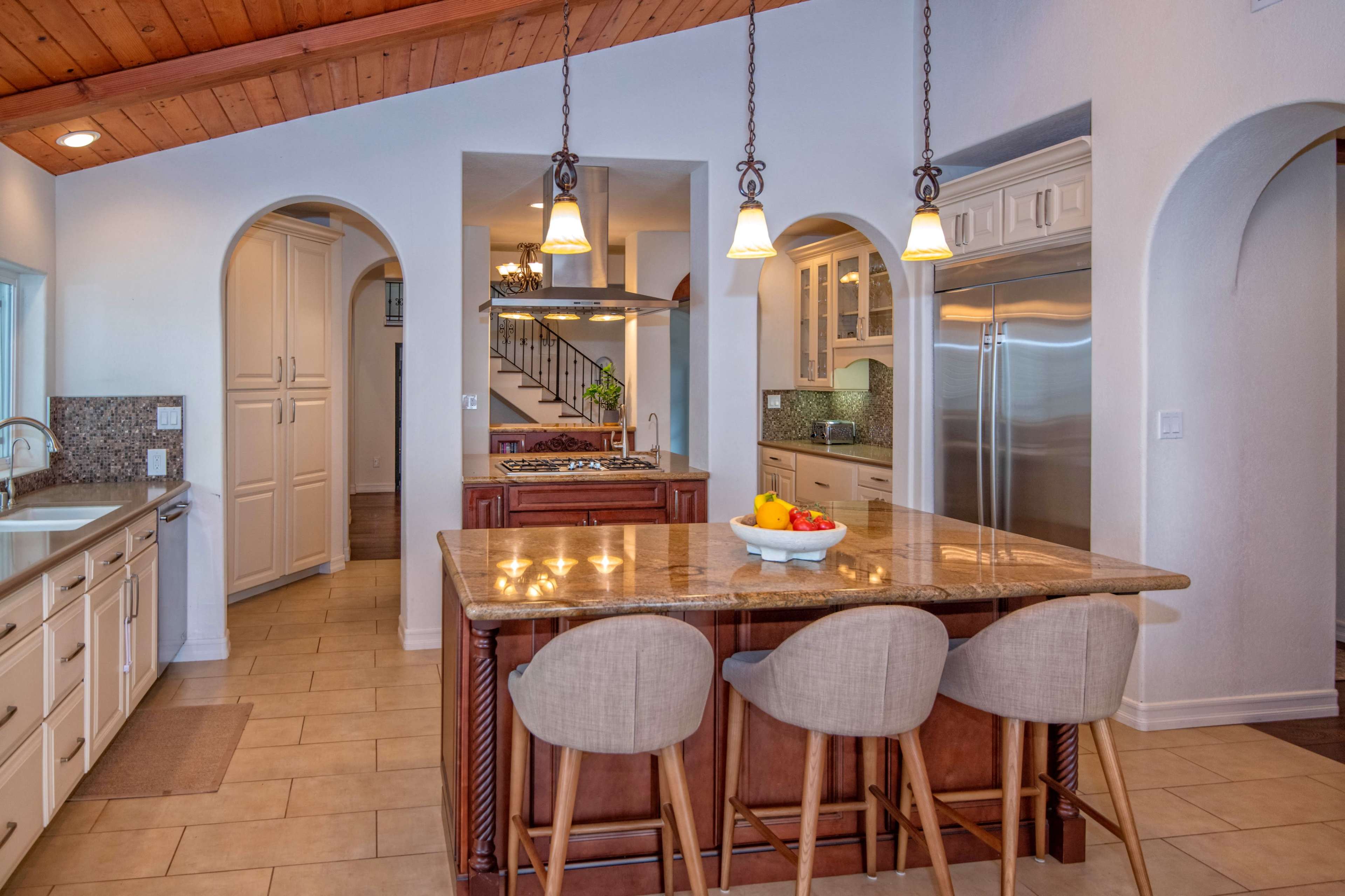 A kitchen with a large island bar featuring three stools, surrounded by cabinetry and appliances, with archways leading to other areas of the home.