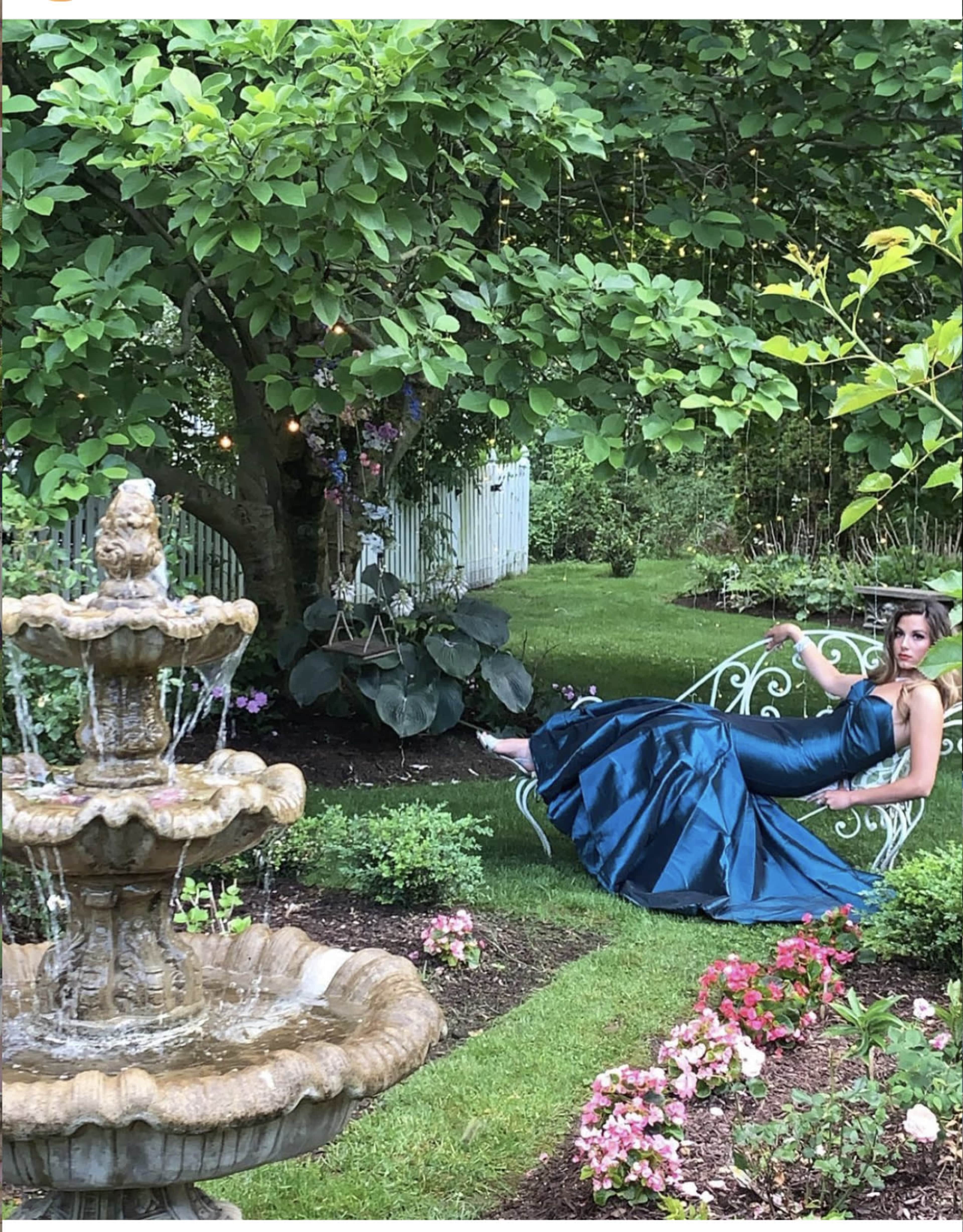 A woman in a blue evening gown relaxes on a white wrought-iron bench in a lush garden with a fountain and colorful flowers.