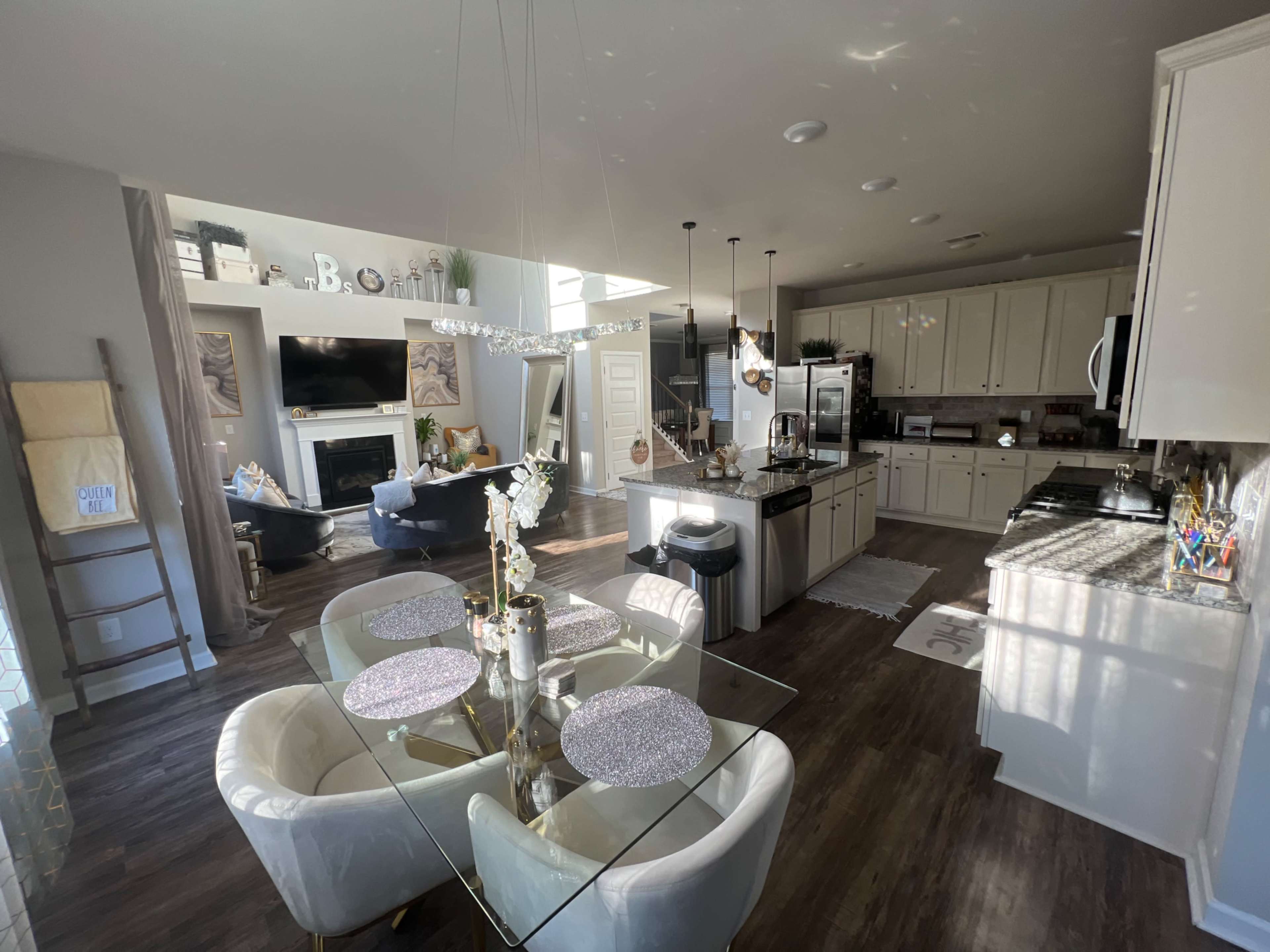 A modern kitchen and dining area with a glass table, light-colored cabinets, and a living room visible in the background.