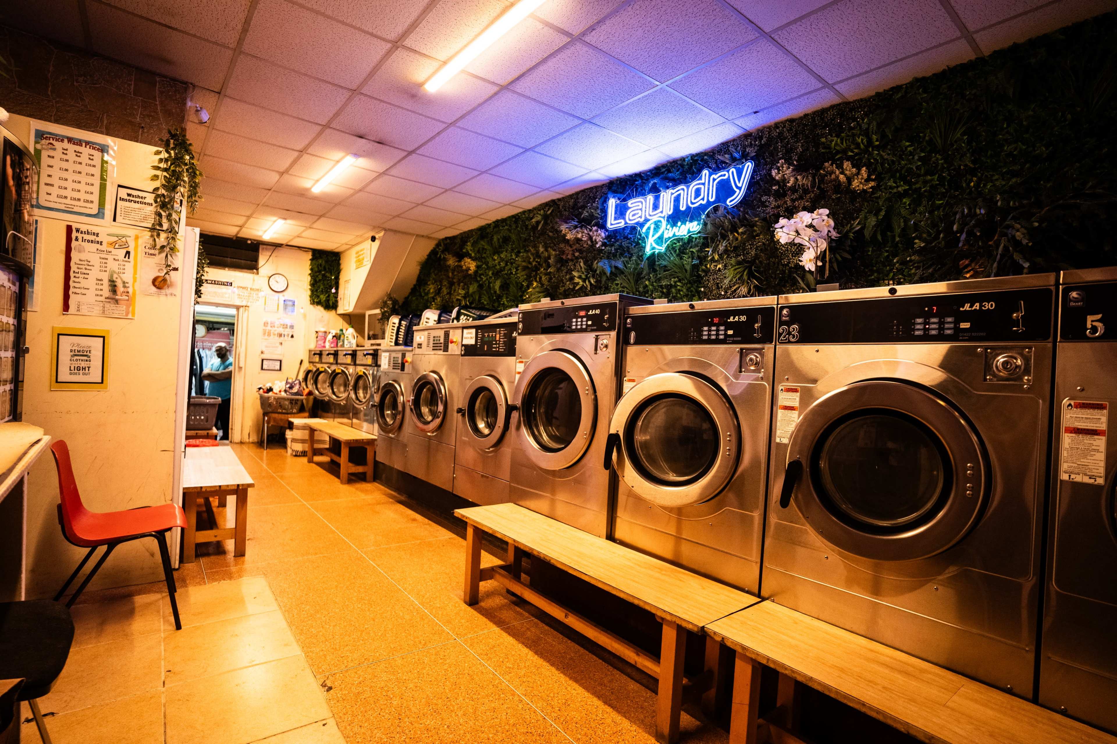 The image shows a row of washing machines in a laundromat, with neon signage and greenery decor overhead.