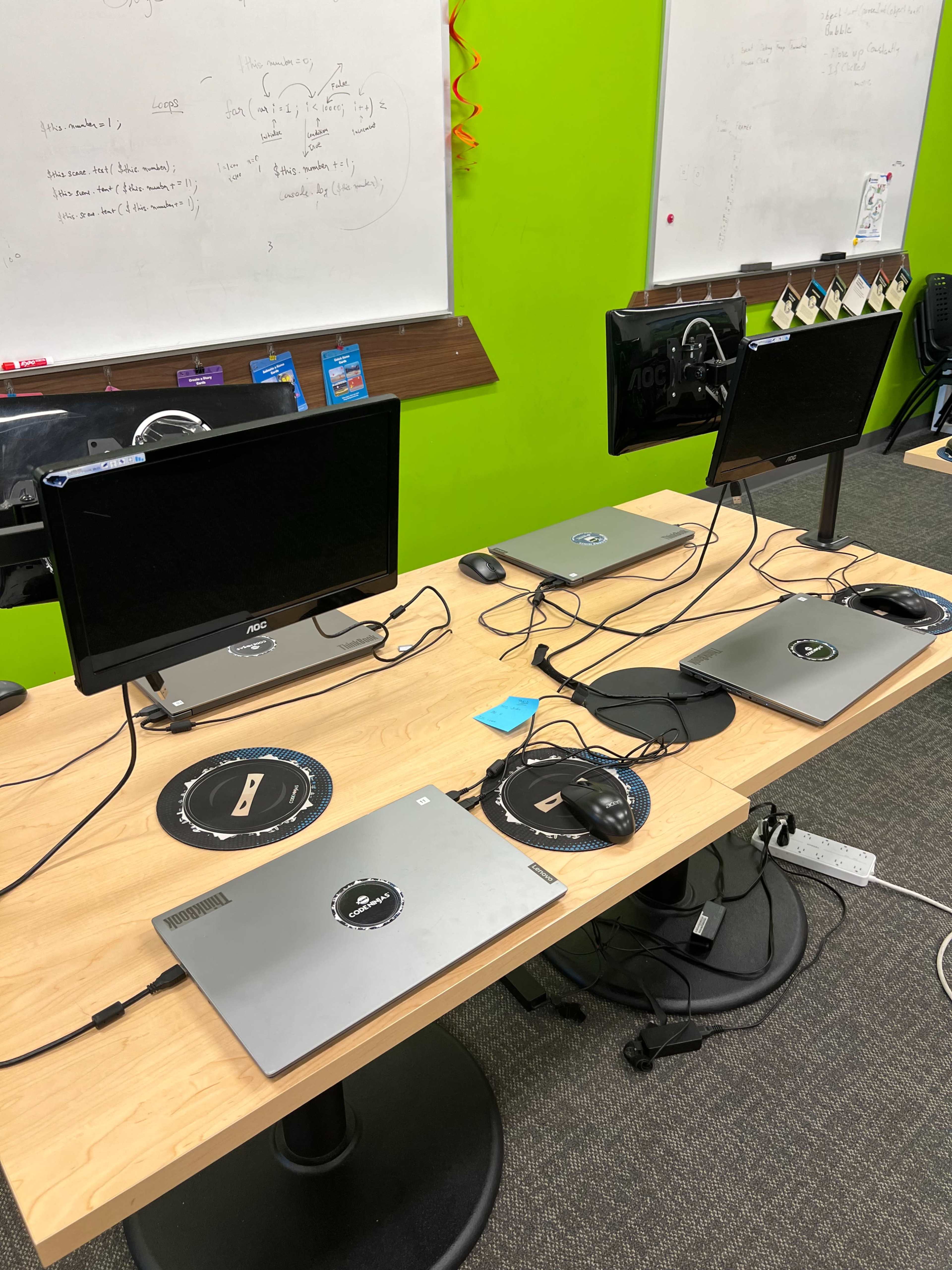 A workspace setup features four laptops and two monitors on a wooden table, with tangled cables and office supplies scattered around.