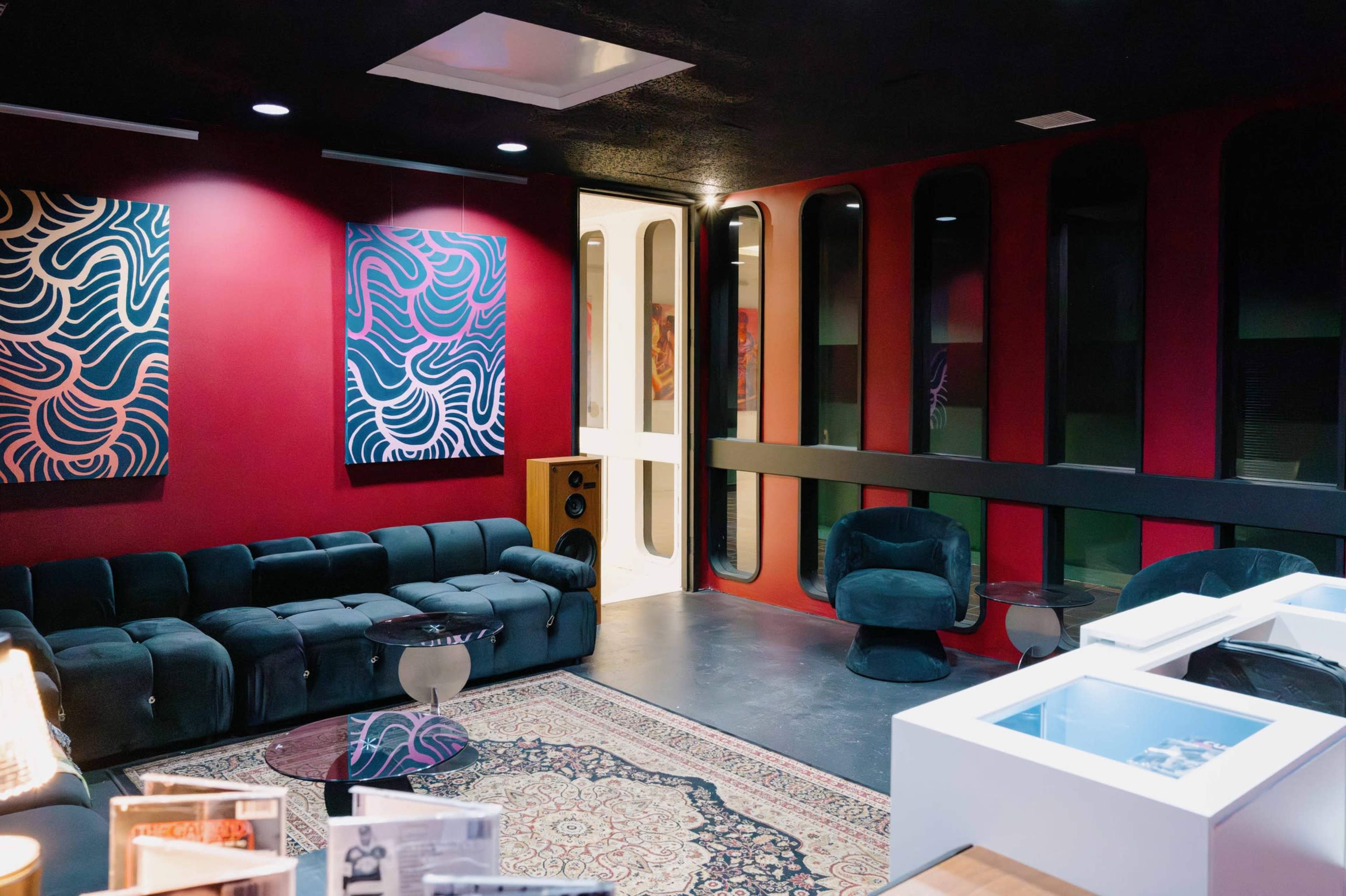 The image shows a modern lounge area featuring a dark red wall, a green velvet sofa, and abstract artwork on display.