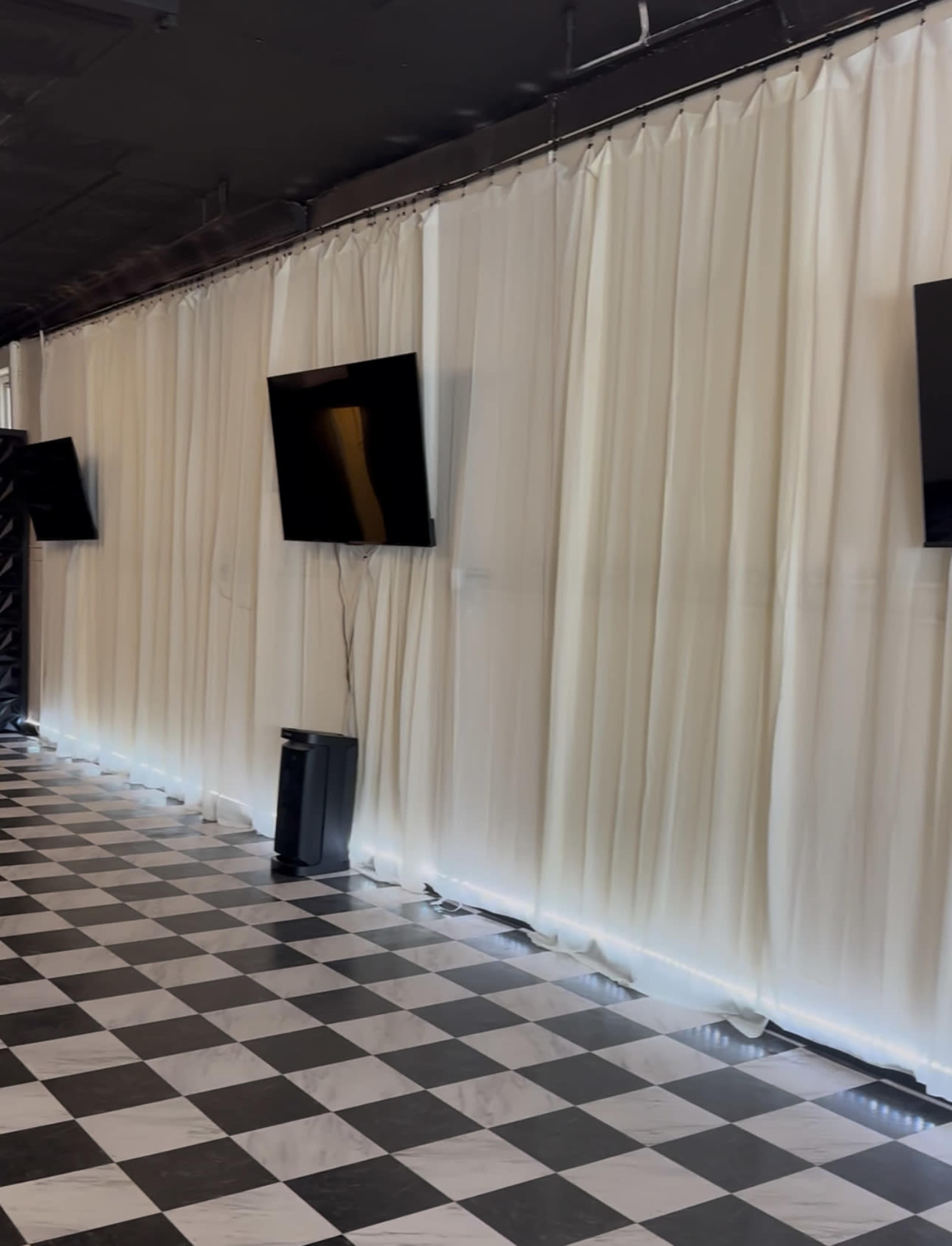The image shows a room with black and white checkered flooring, white draped curtains, and two mounted flat-screen TVs on the walls.