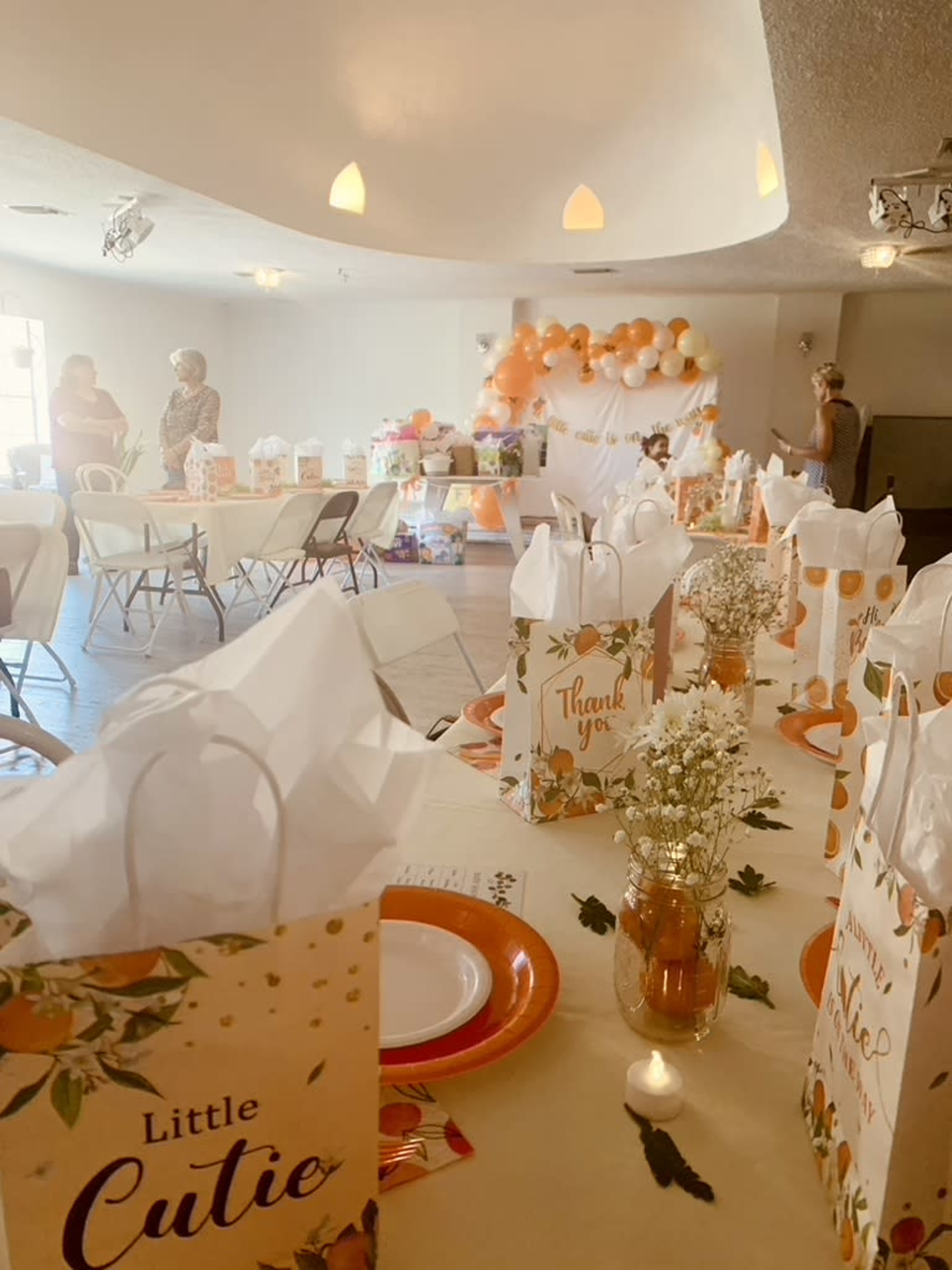 A decorated indoor space features tables set with gift bags and food, while balloons and a dessert table are visible in the background.